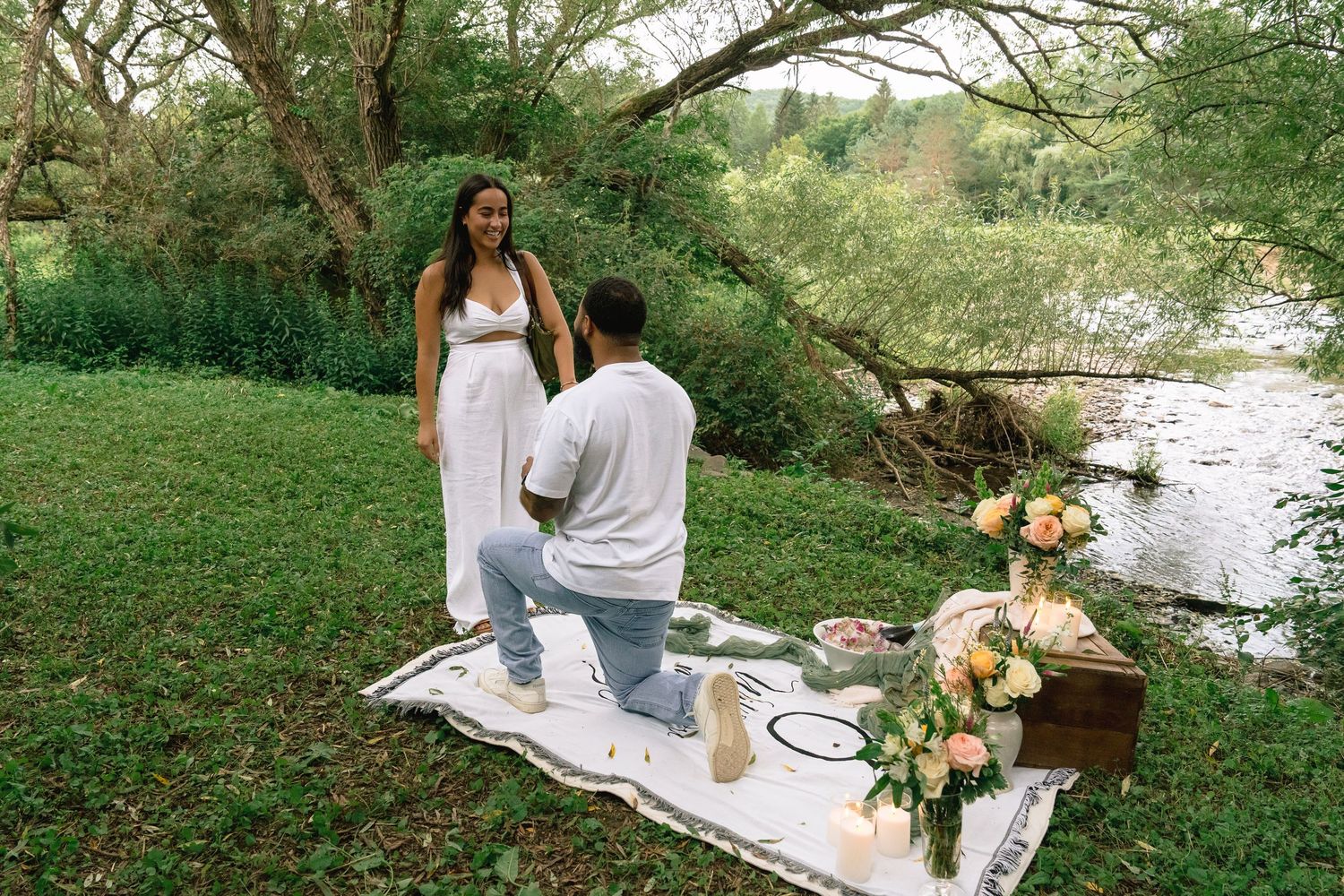 A romantic picnic setup by a peaceful stream with a white blanket and colorful floral bouquet in warm sunlight.