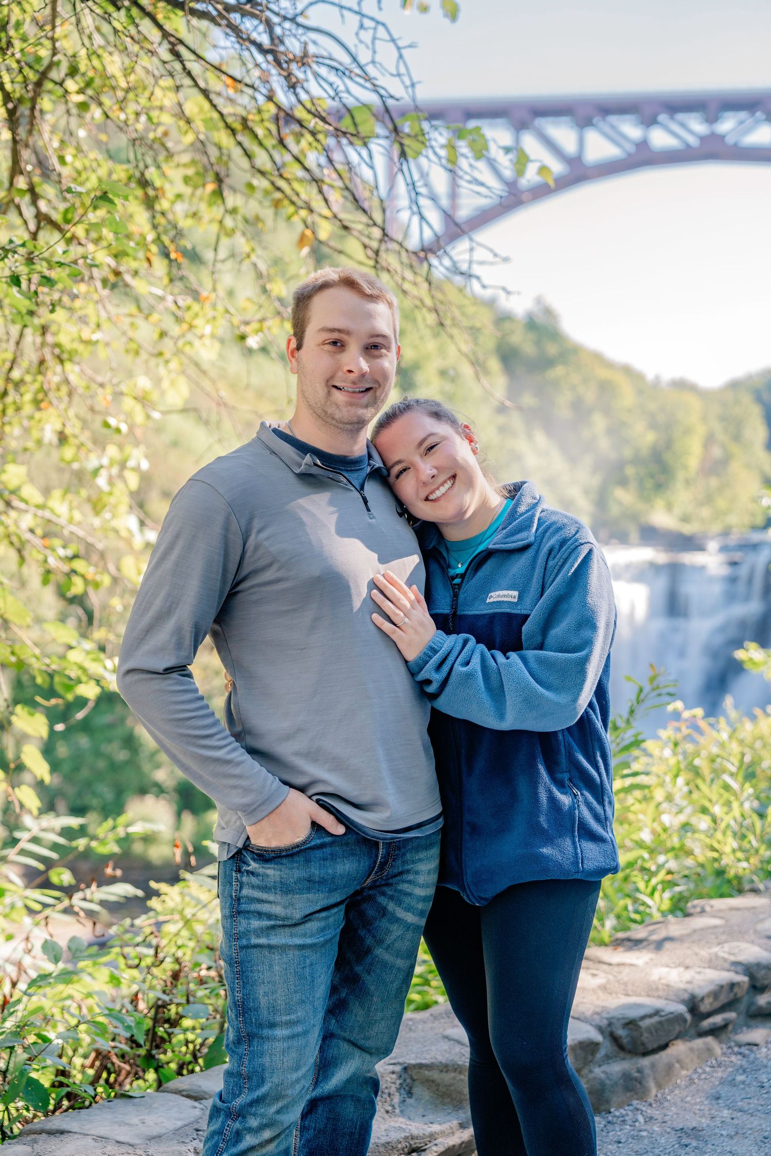 A couple embraces and smiles while posing together outdoors on a sunny autumn day with trees in the background.
