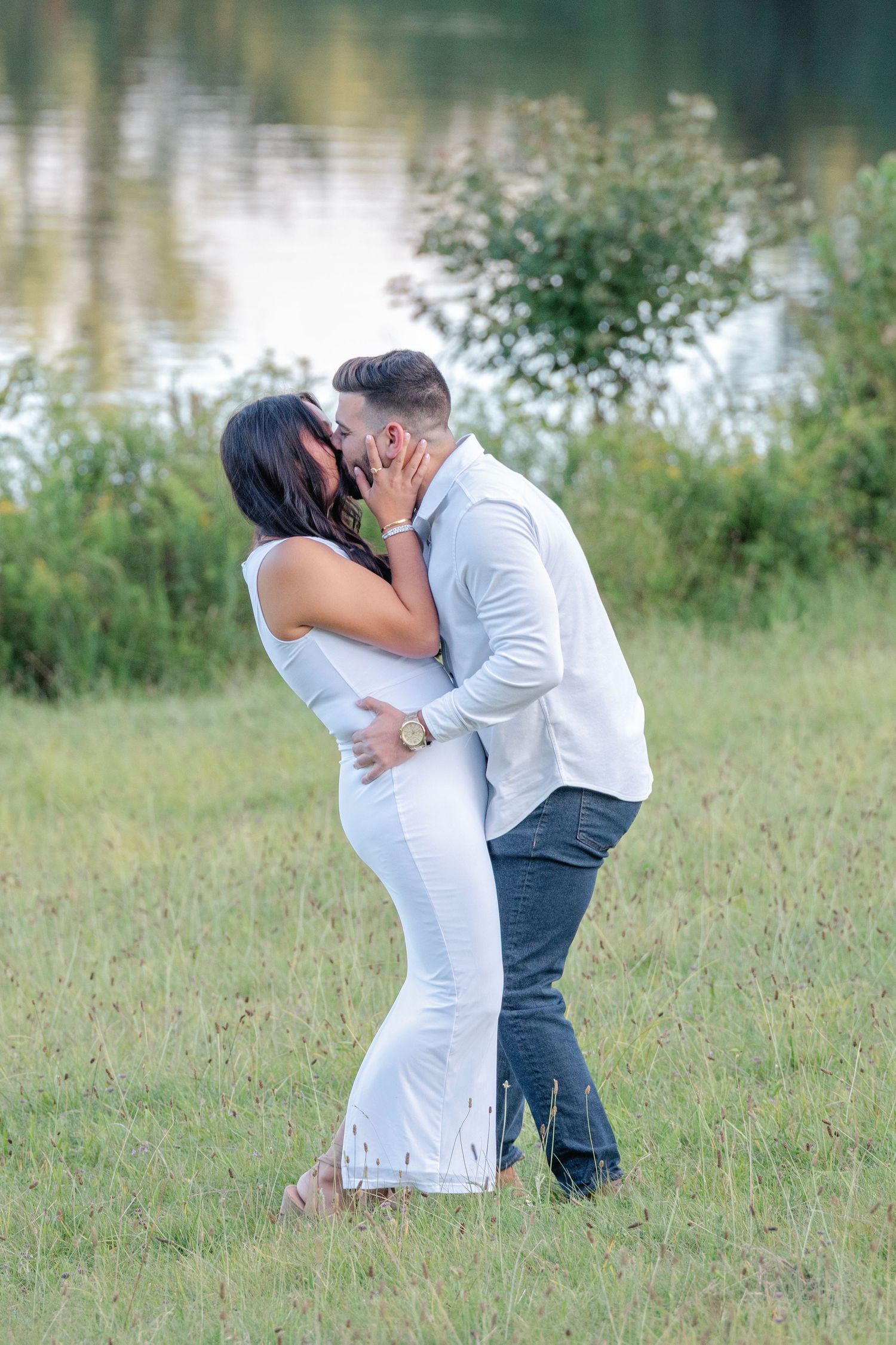 A romantic couple in white and grey attire share intimate moments by a lakeside on a grassy field.