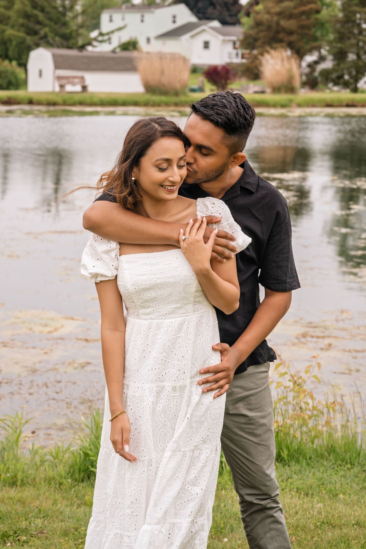 A romantic couple embraces by a serene lakeside, with the woman wearing a flowing white dress and the man in casual attire.
