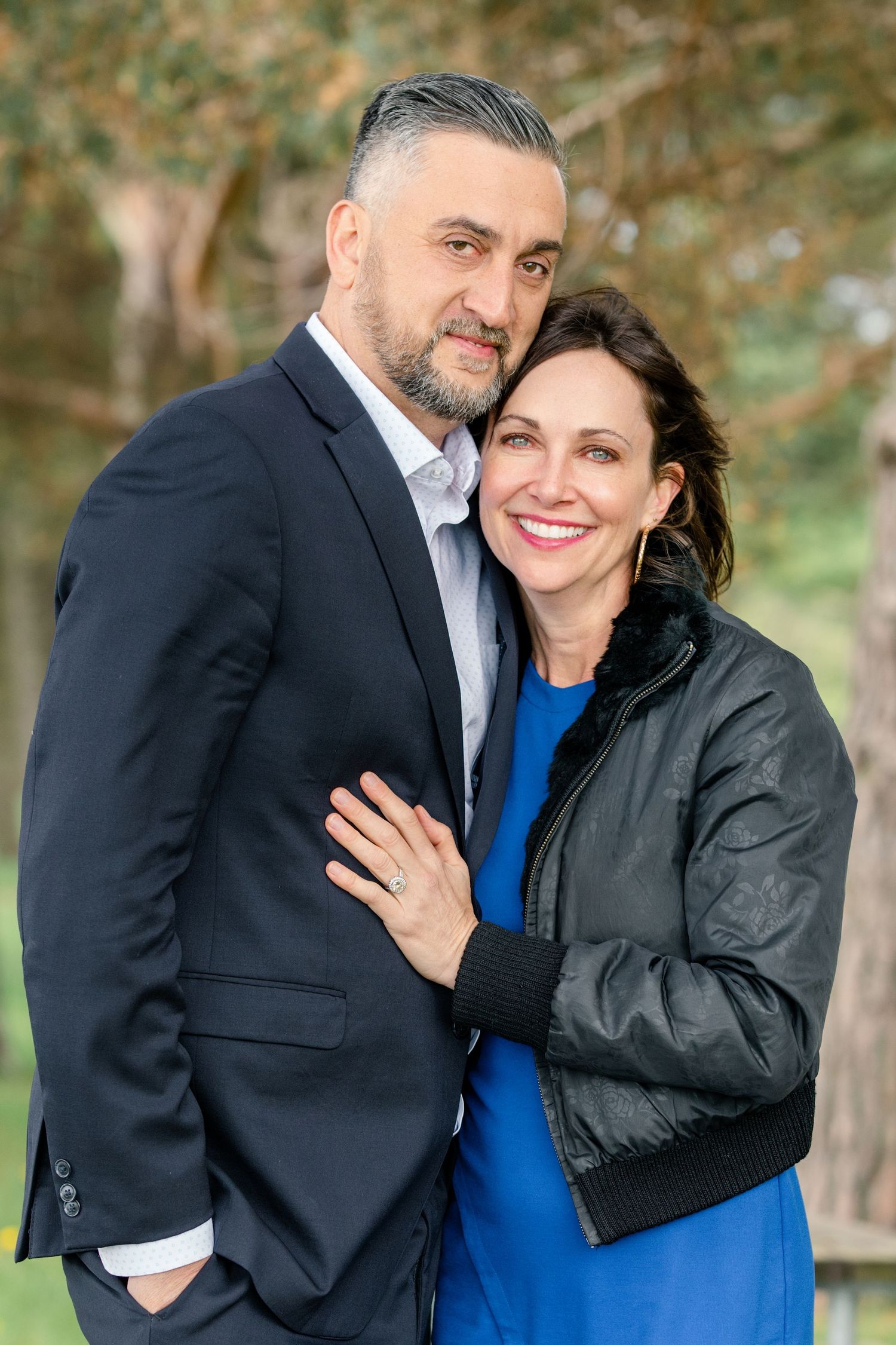 A romantic outdoor photoshoot showing a couple embracing and smiling together against a backdrop of trees.
