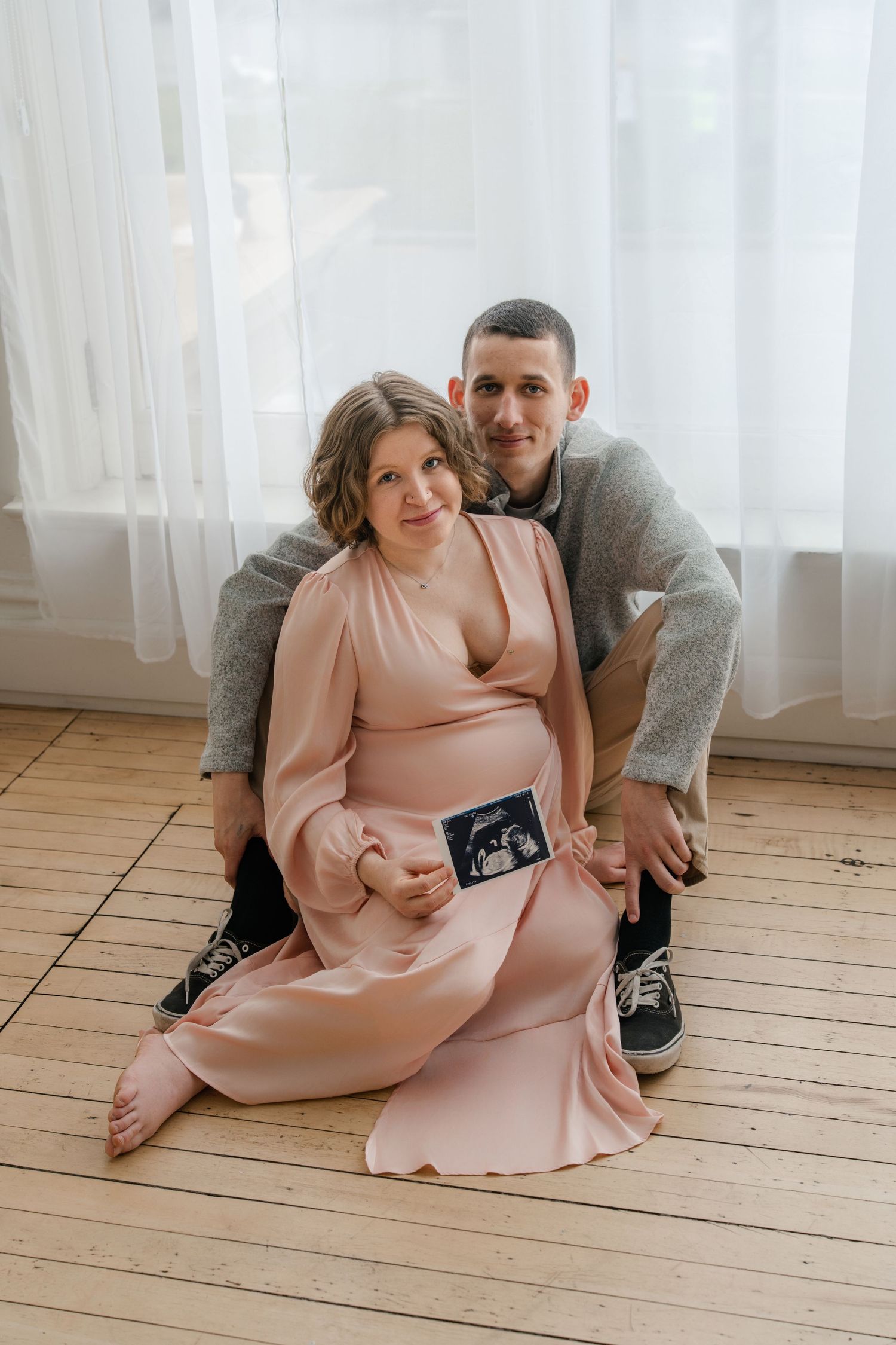 A couple sits on wooden flooring by sheer curtains while holding an ultrasound photo in a pregnancy announcement pose.
