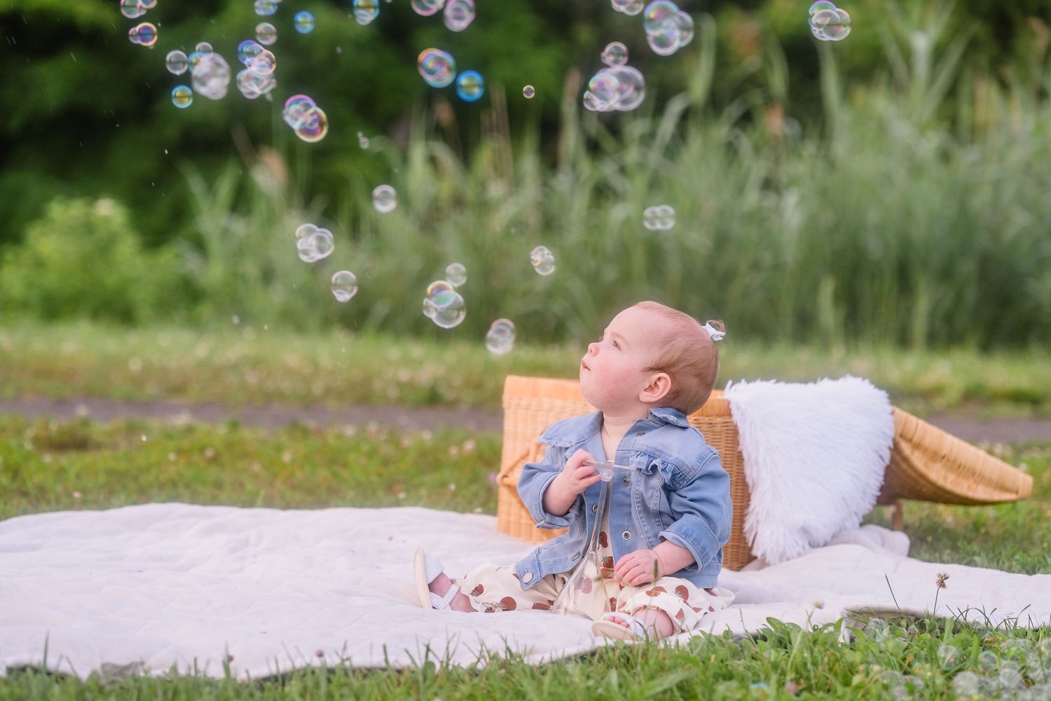 A baby in denim lying on a white blanket outdoors while soap bubbles float through the air on a sunny day.