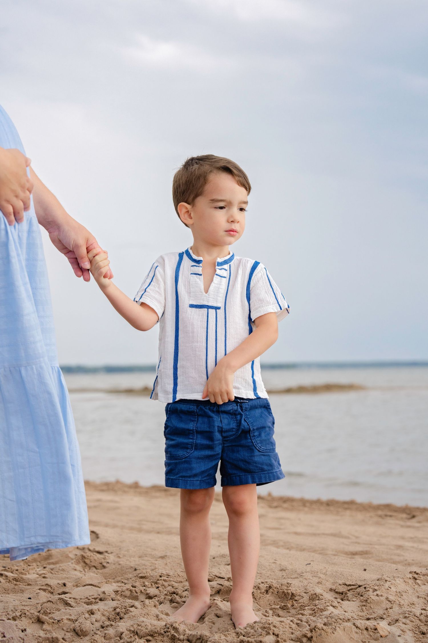 Young child in striped shirt and blue shorts holding hands with someone while standing barefoot on a beach.