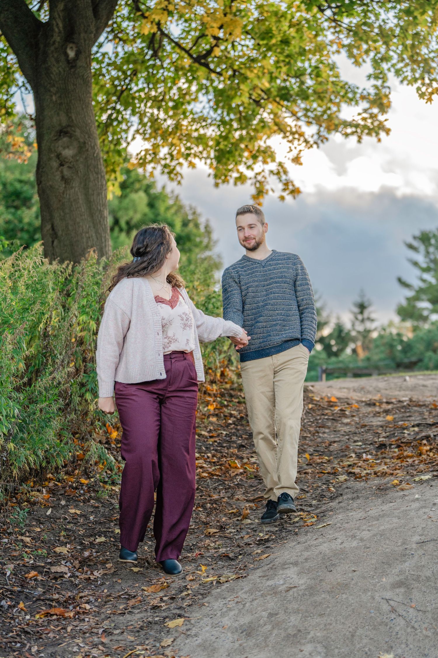 A couple walks along a leaf-covered autumn path under golden trees during a romantic outdoor photoshoot.