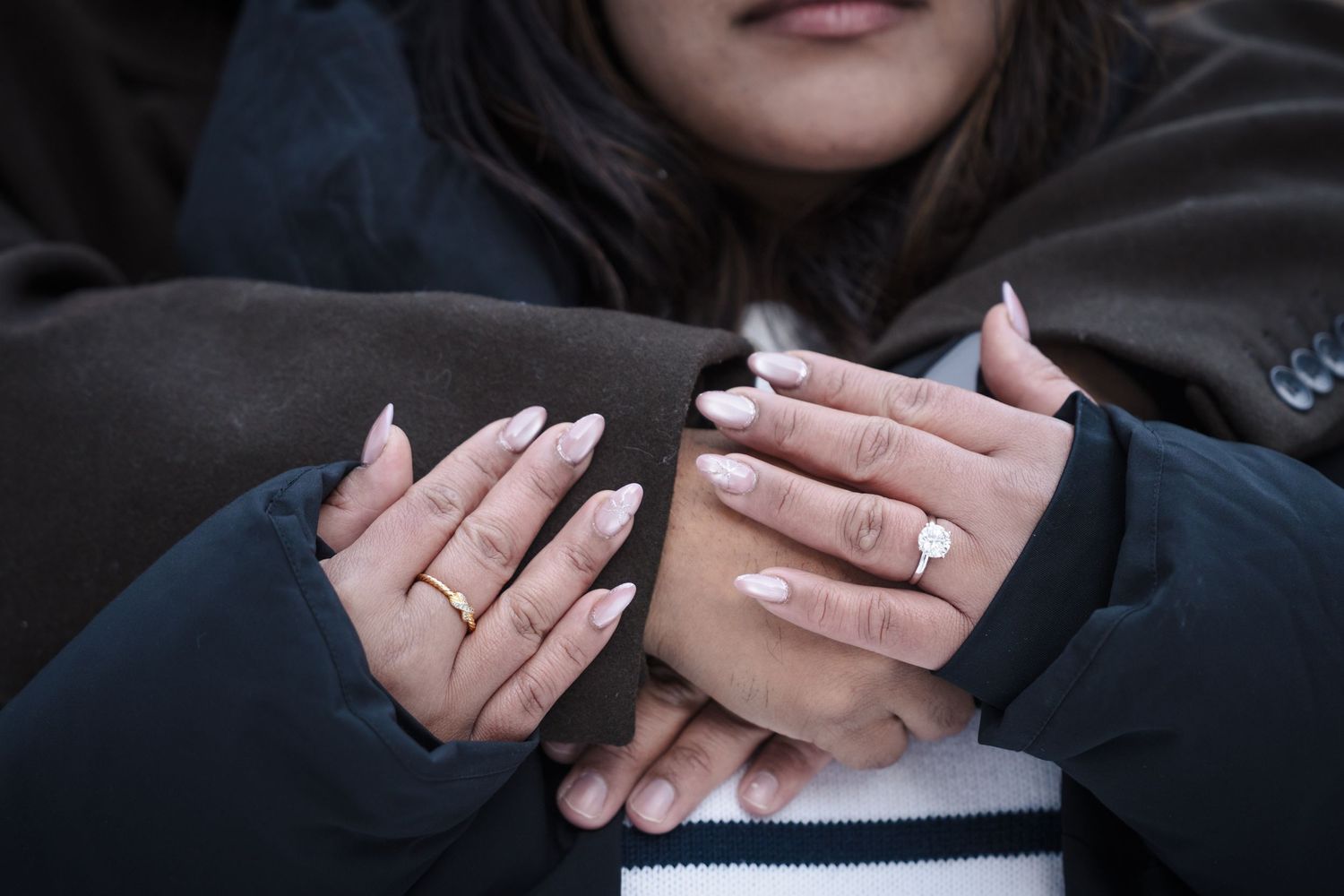 Close up view of hands with engagement ring and manicured nails against dark background.