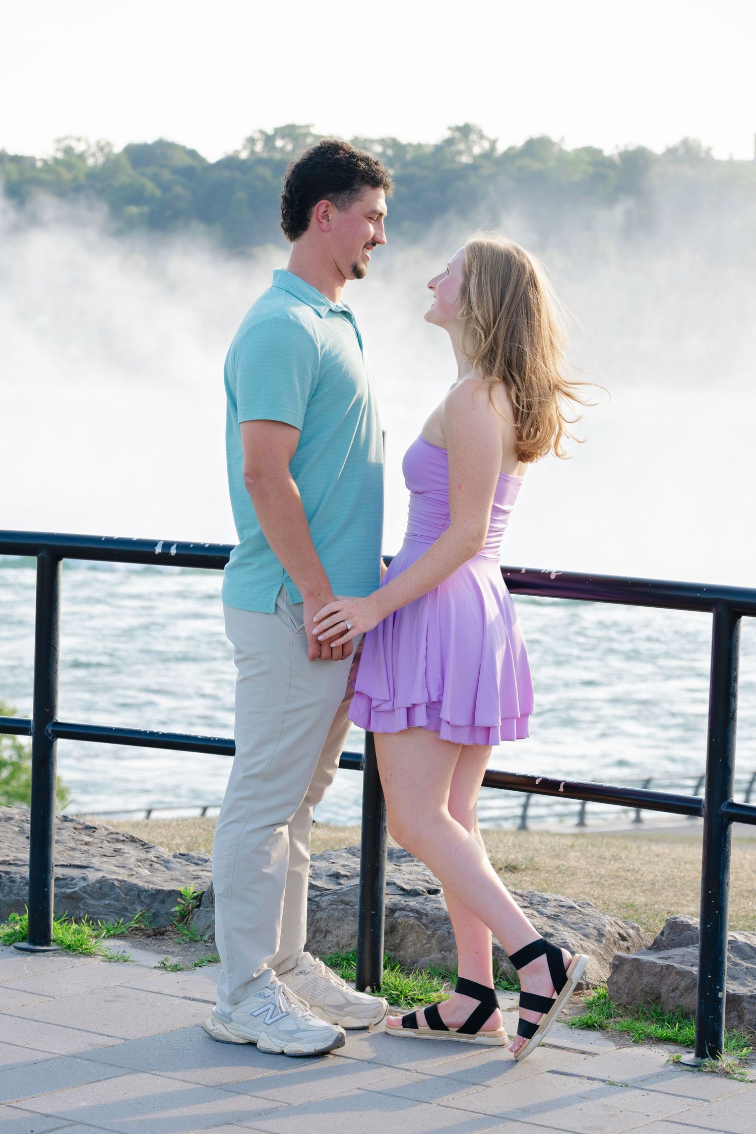 A romantic couple poses together on a misty morning by the water, with her wearing a purple dress and him in blue.