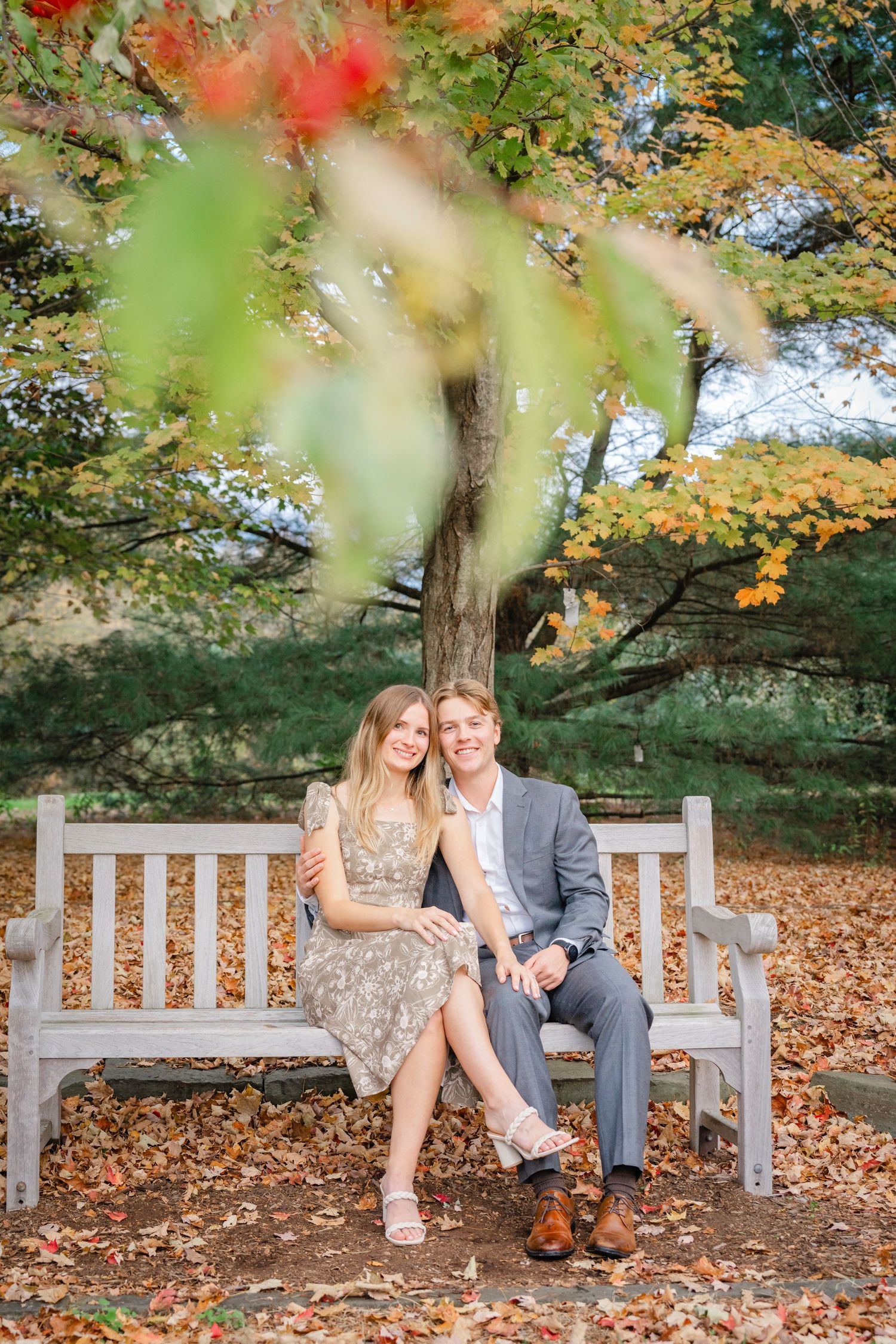 A couple sits together on a wooden bench surrounded by fallen autumn leaves and colorful fall foliage in a park setting.