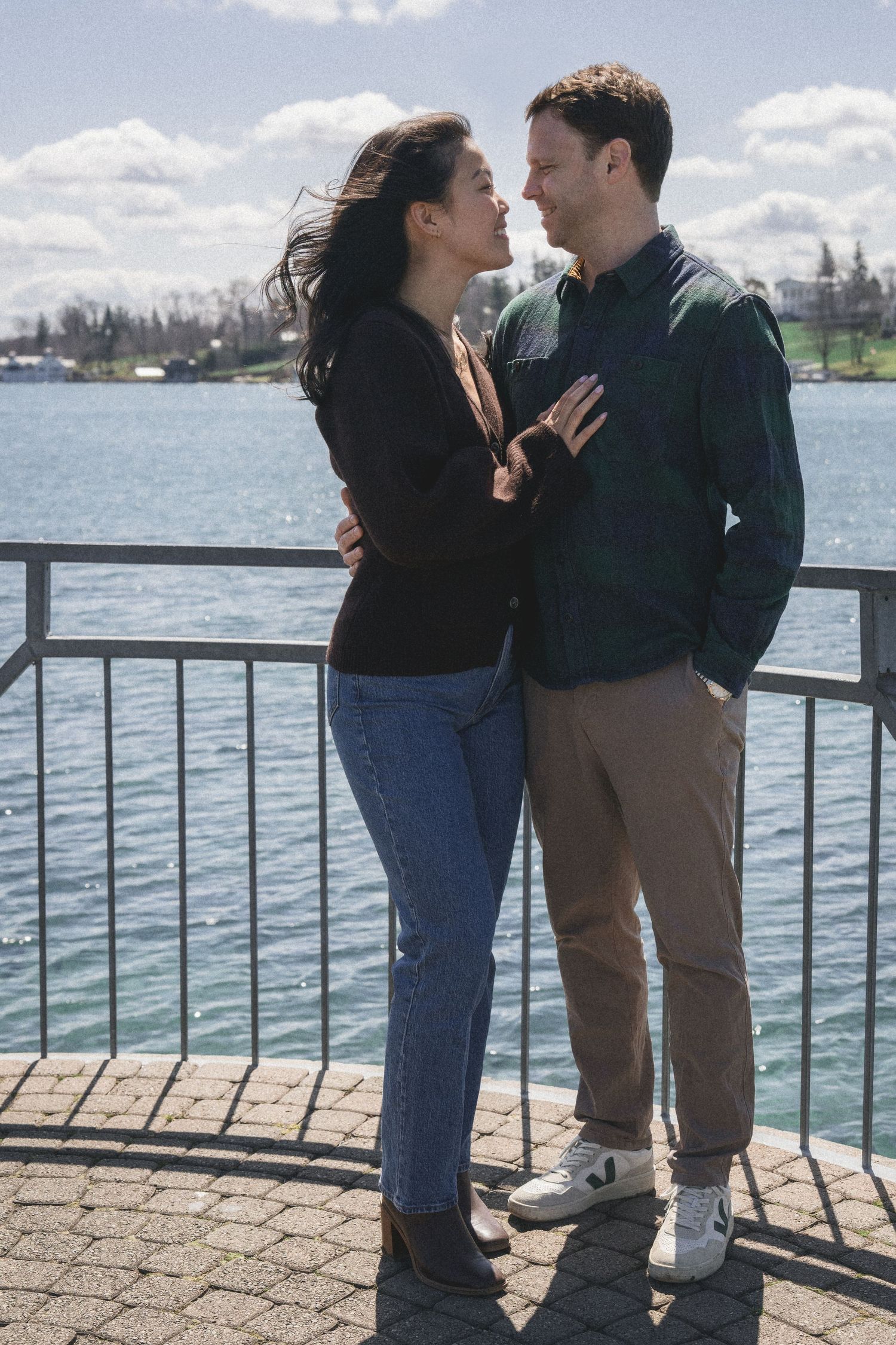A couple sharing intimate moments on a waterfront pier with blue water and clouds in the background.