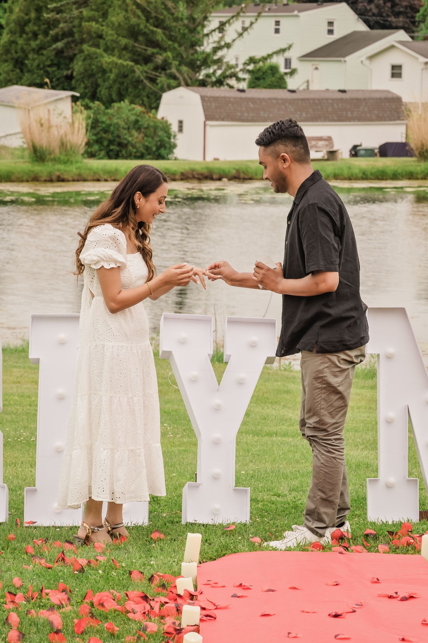 A romantic proposal setup with red blanket and white letter decorations by a lake.
