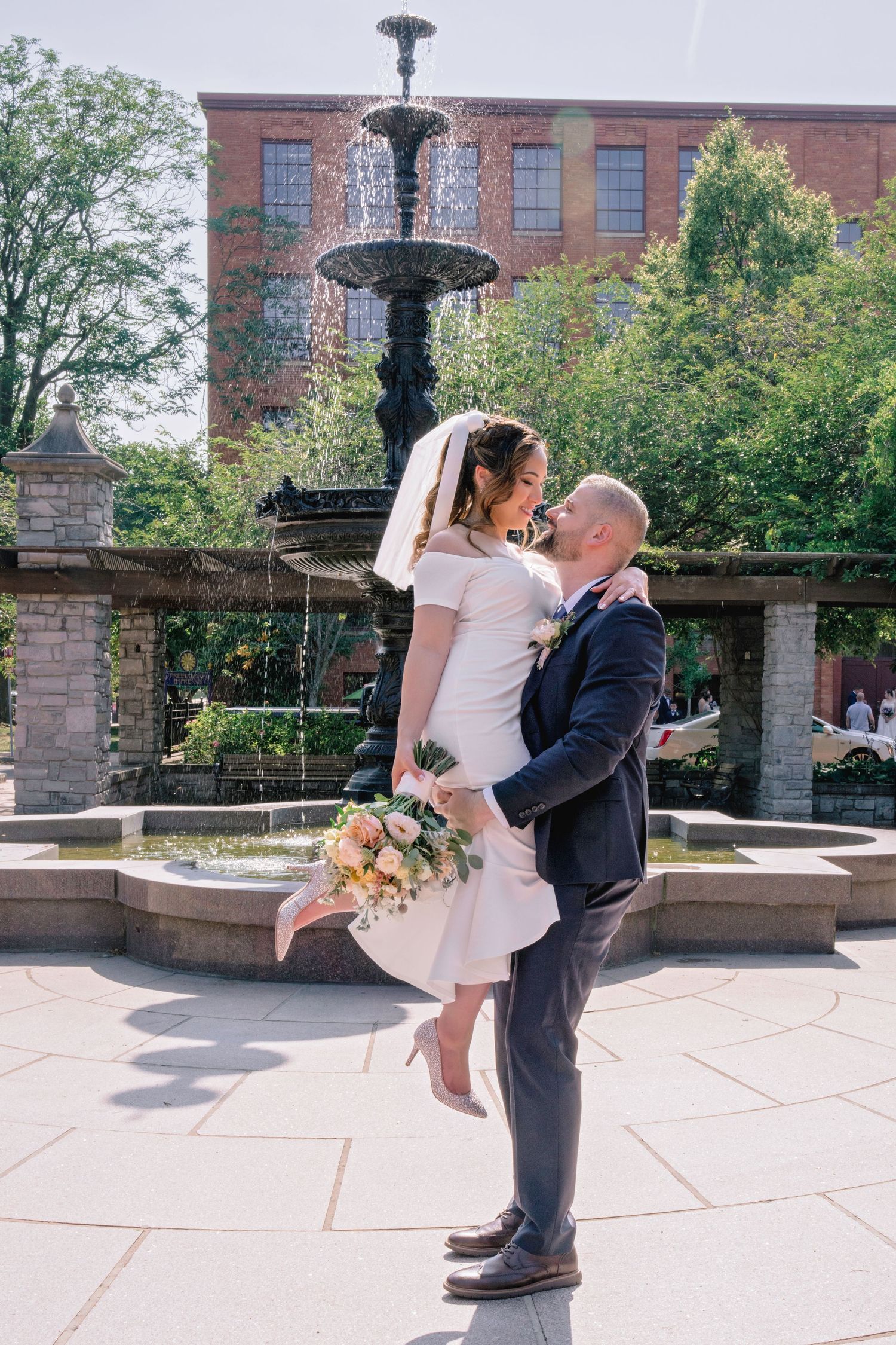 Groom lifts bride in white dress and veil near ornate fountain, holding bouquet of pink roses outdoors.