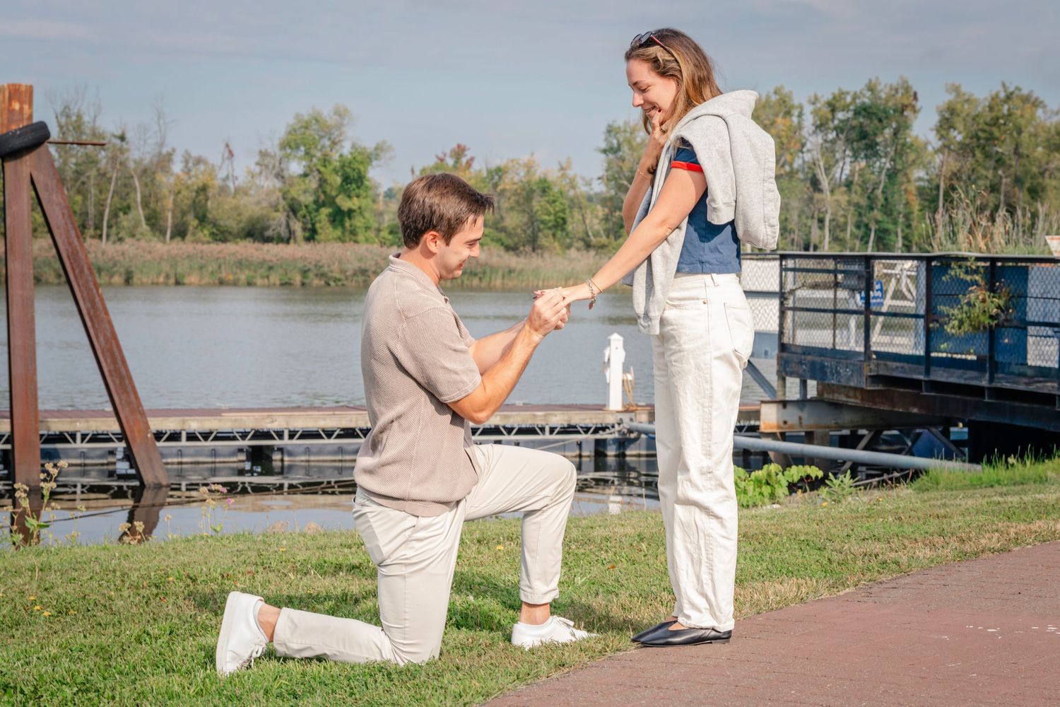 A romantic marriage proposal takes place on a scenic dock by a lake on a sunny afternoon.