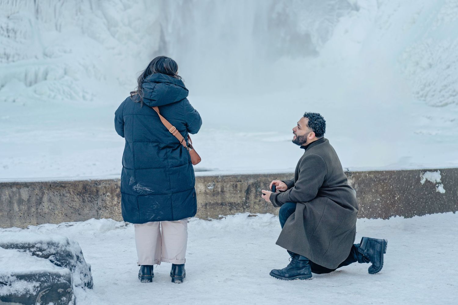 Two people crouch in the snow while taking photographs near a snowy wall.