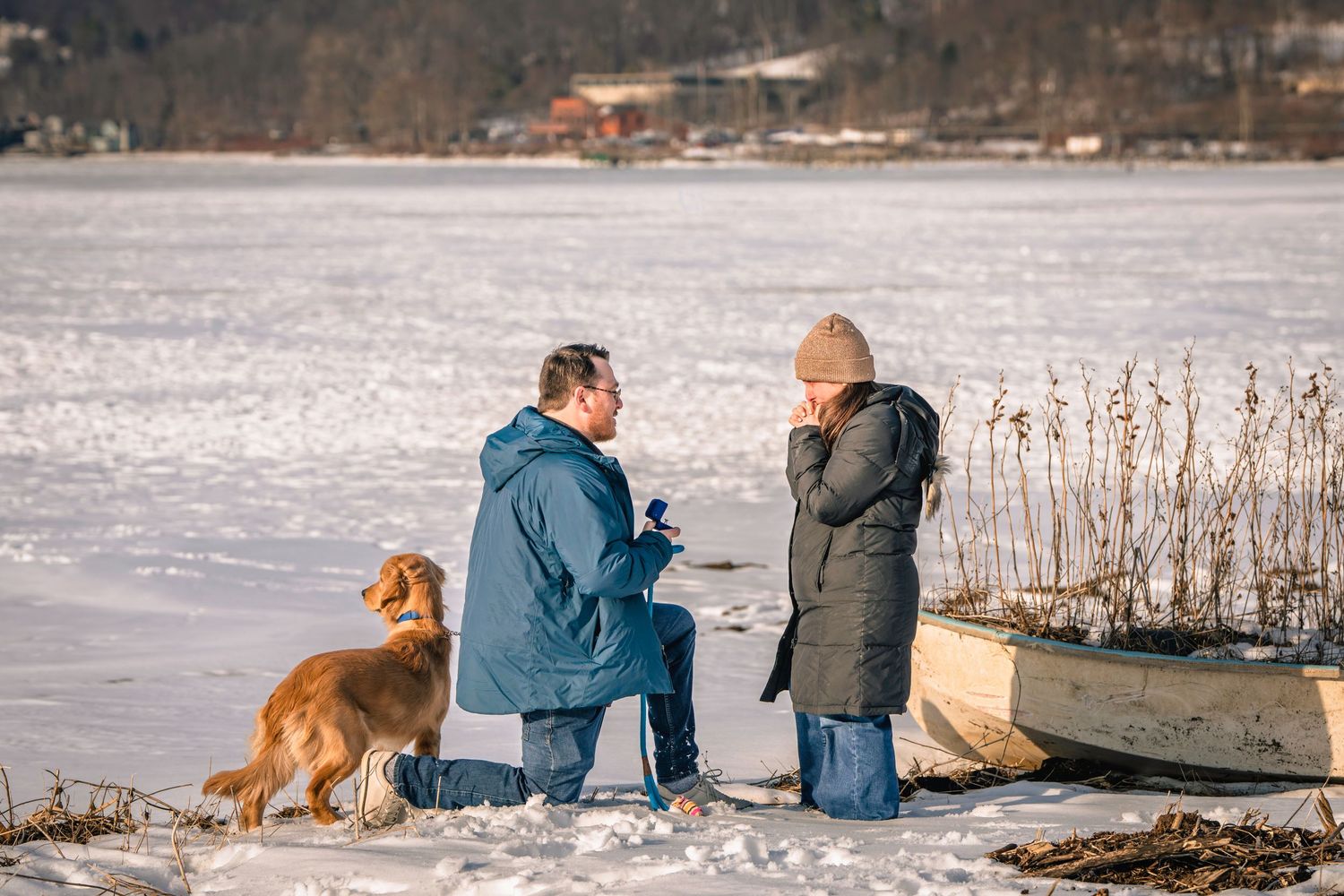 Couple sharing a marriage proposal moment by frozen lake in winter with golden retriever dog nearby.