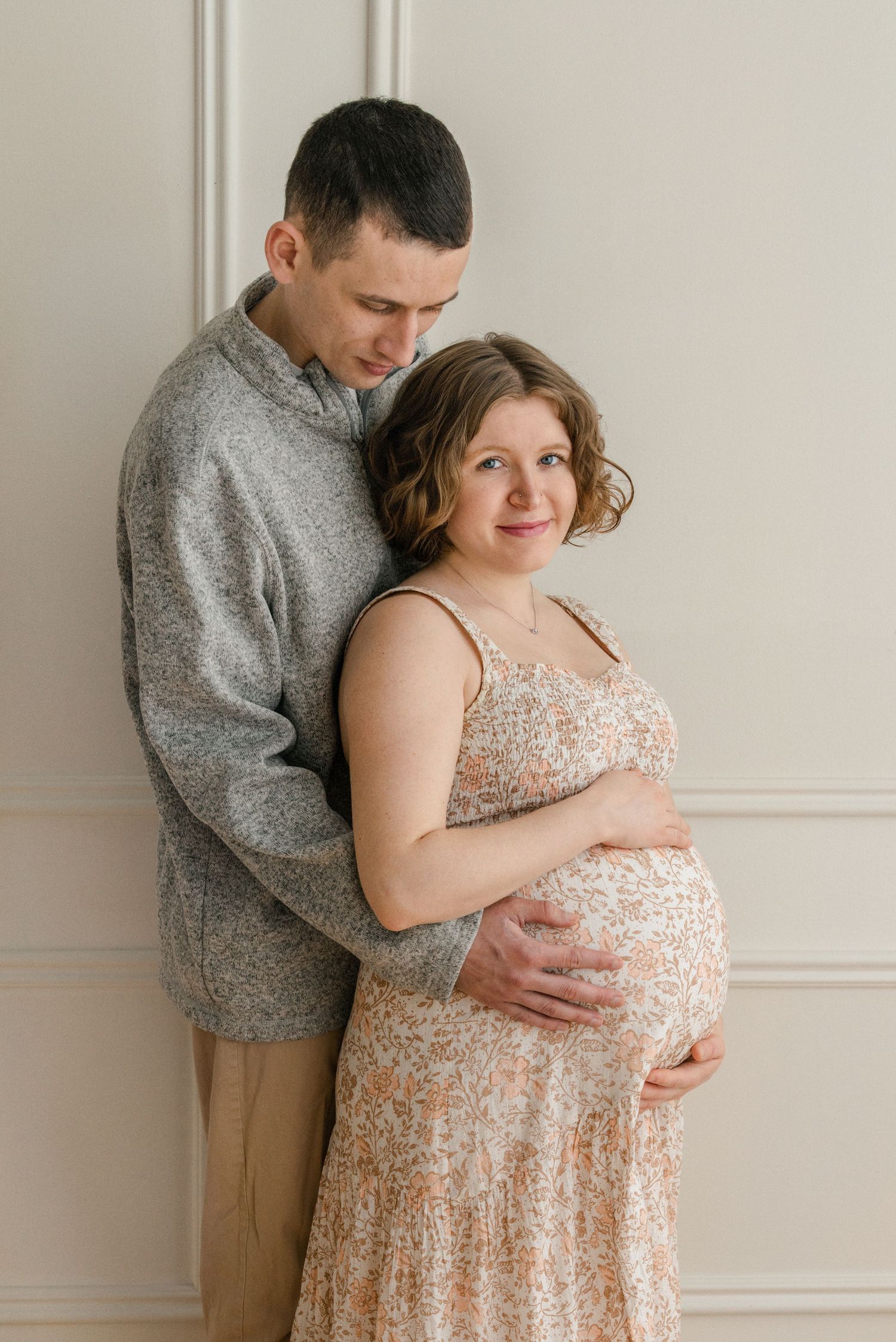 A tender maternity photo shows a couple embracing against a light wall, with her wearing a sparkly blush-colored dress.