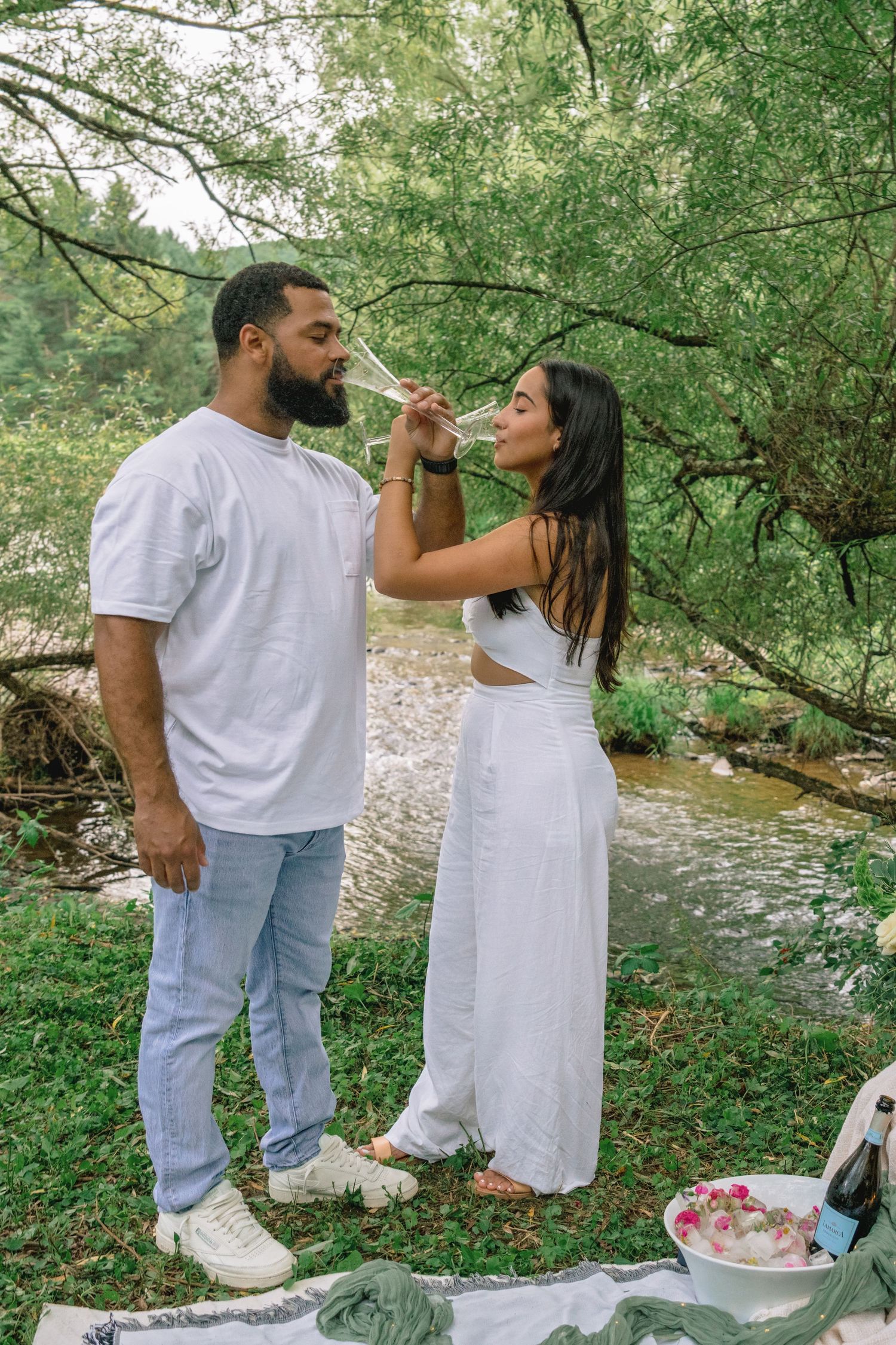 A couple enjoys a romantic picnic by a stream in a lush green forest setting.