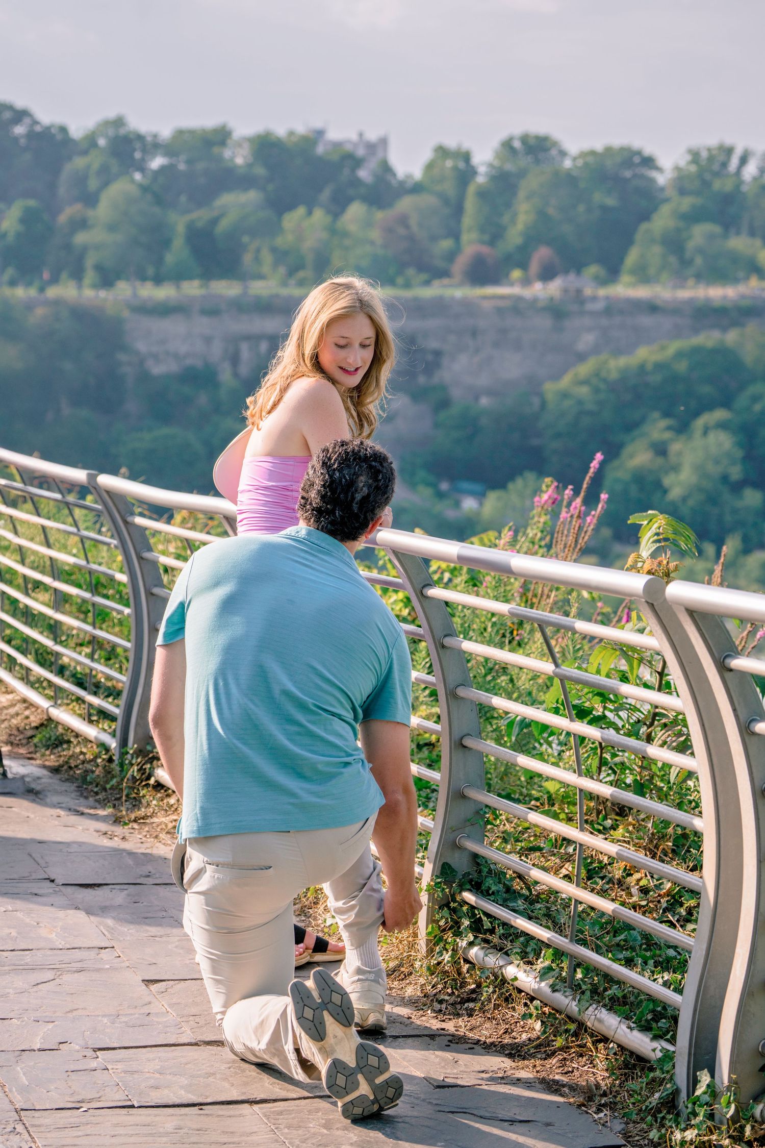 A couple shares a romantic moment on a scenic overlook with metal railings and greenery in the background.