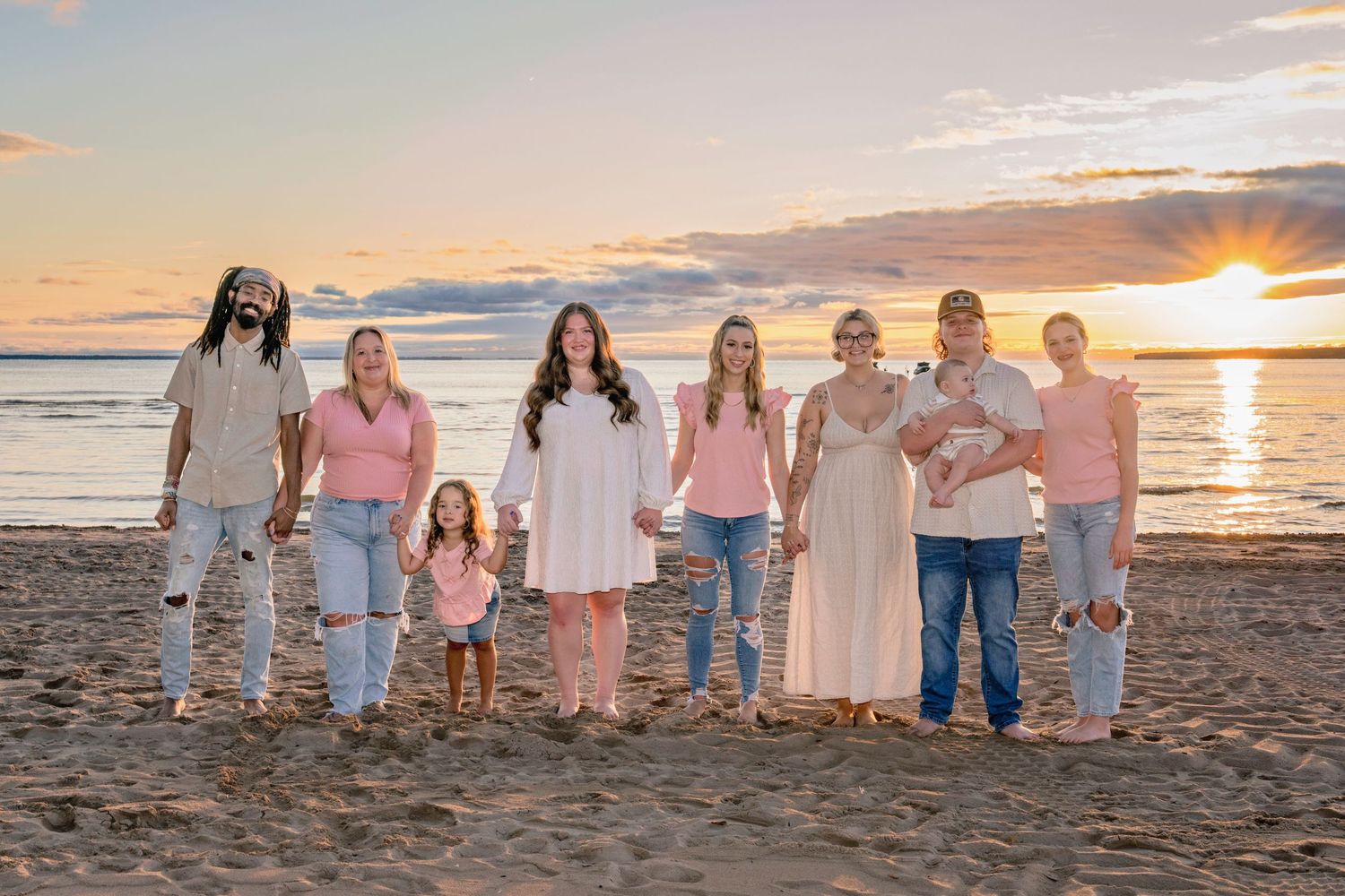Extended family group poses together on beach at sunset wearing coordinated white and pink outfits.