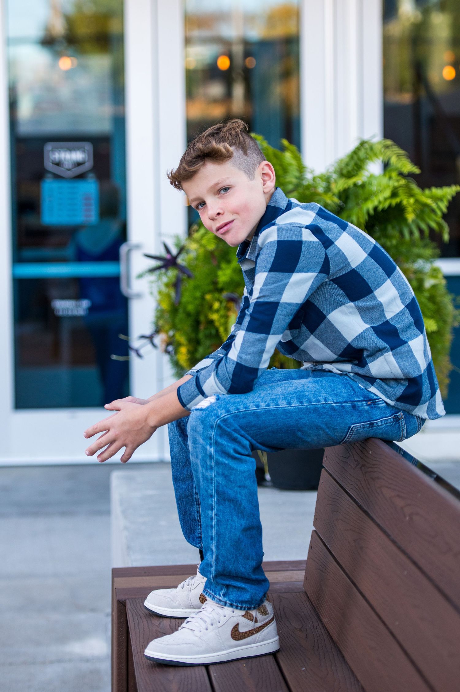 Teen in plaid shirt and jeans sits casually on wooden bench outside storefront downtown Overland Park