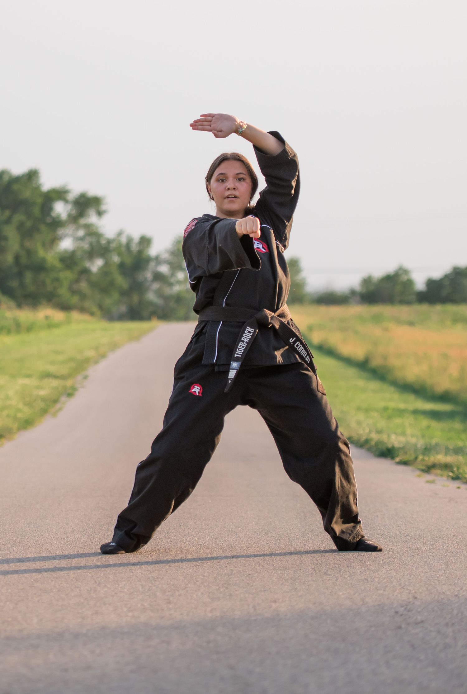 Person in black martial arts uniform performs stance on paved path outdoors.