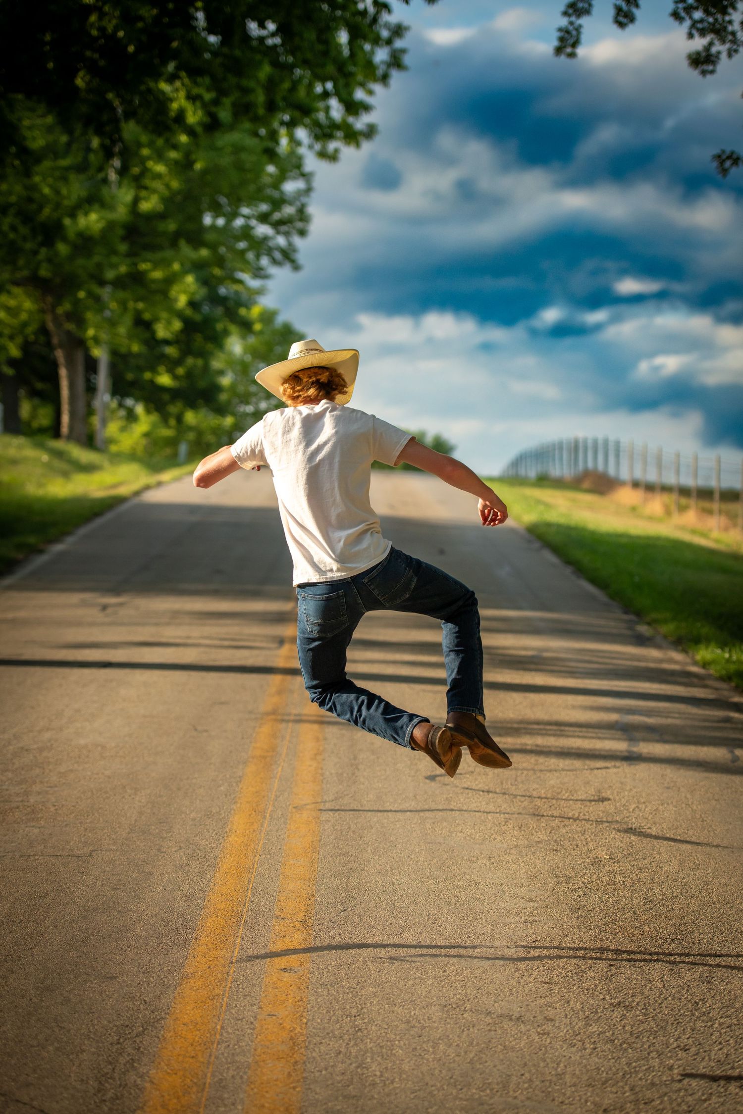 Person in a straw hat jumping playfully on a rural road with blue sky and trees.