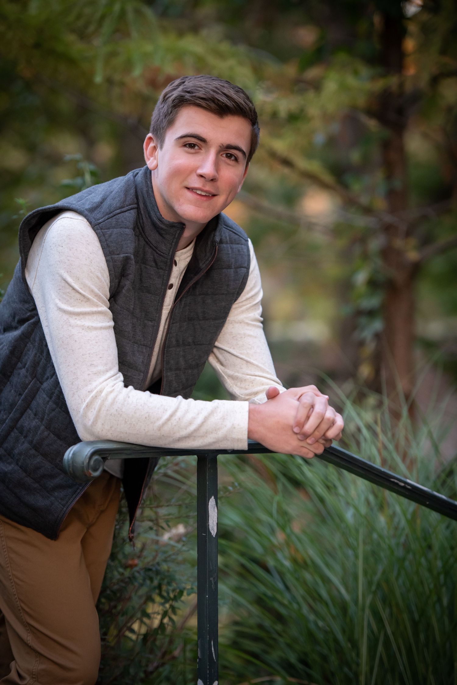 Young person in cream sweater and dark vest poses outdoors leaning on railing in natural setting.
