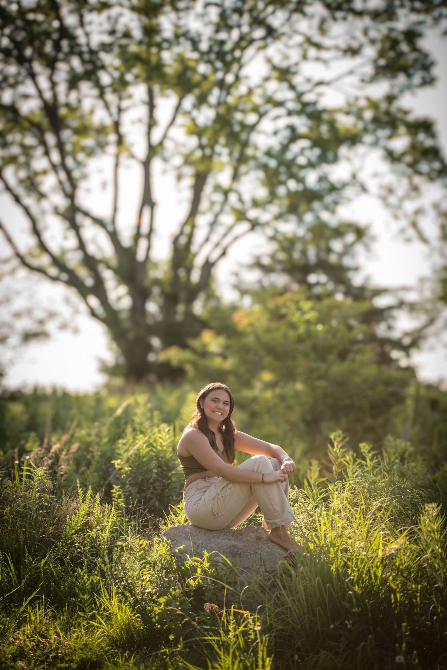Senior sits peacefully on rock surrounded by tall grass during golden hour sunset.