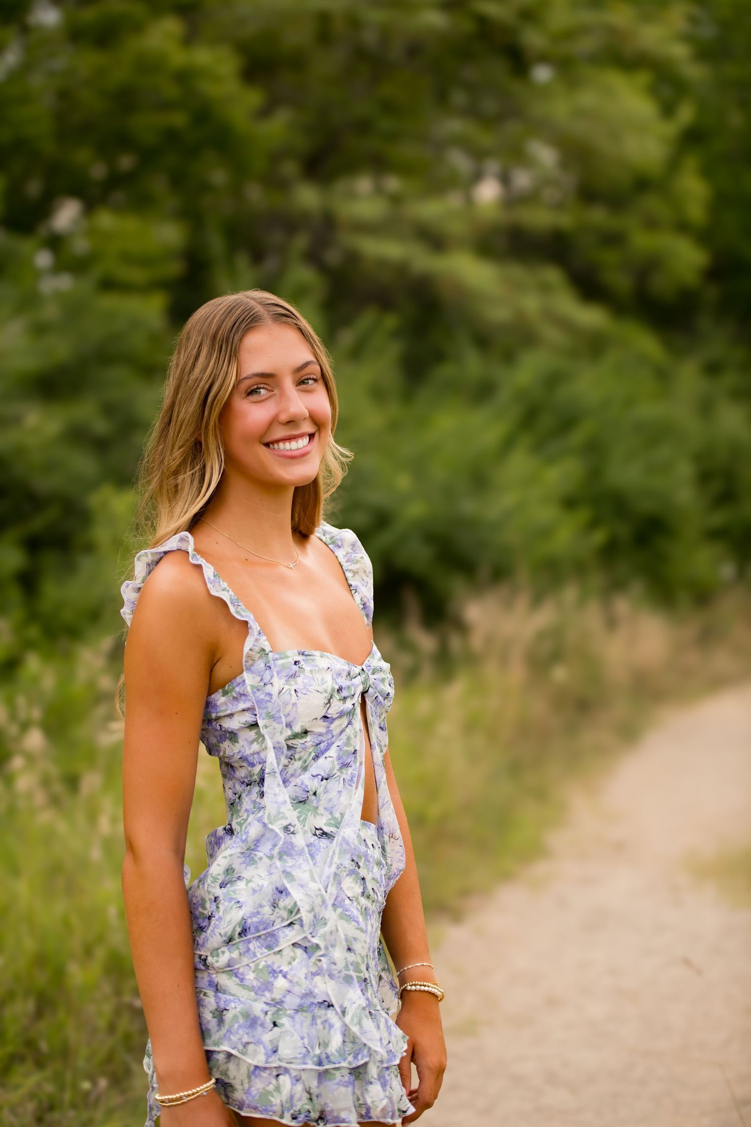 Blue Valley senior in floral summer dress smiles while standing on nature trail with greenery in background.