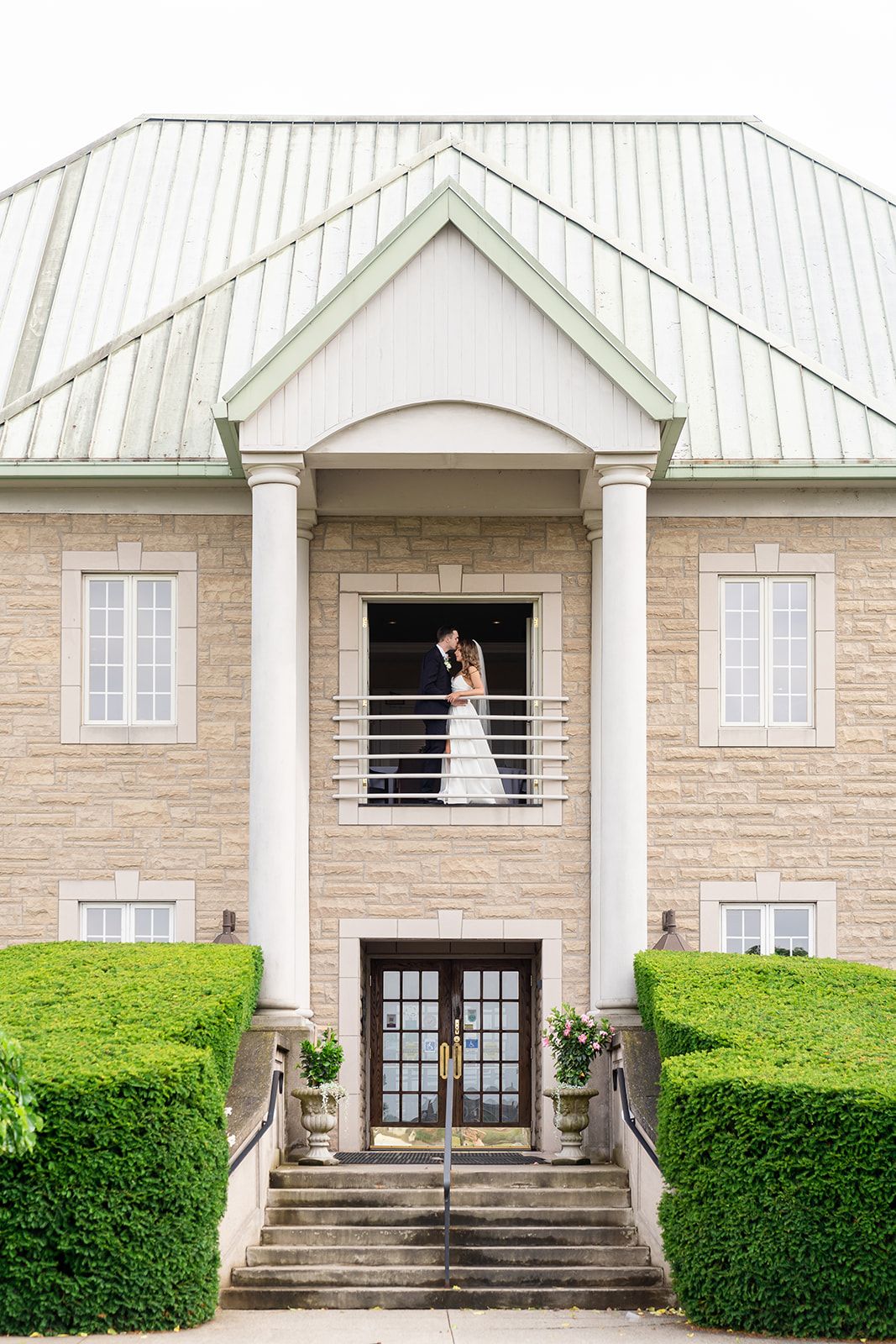 Elegant stone mansion with white trim features a balcony where newlyweds pose against symmetrical architecture.