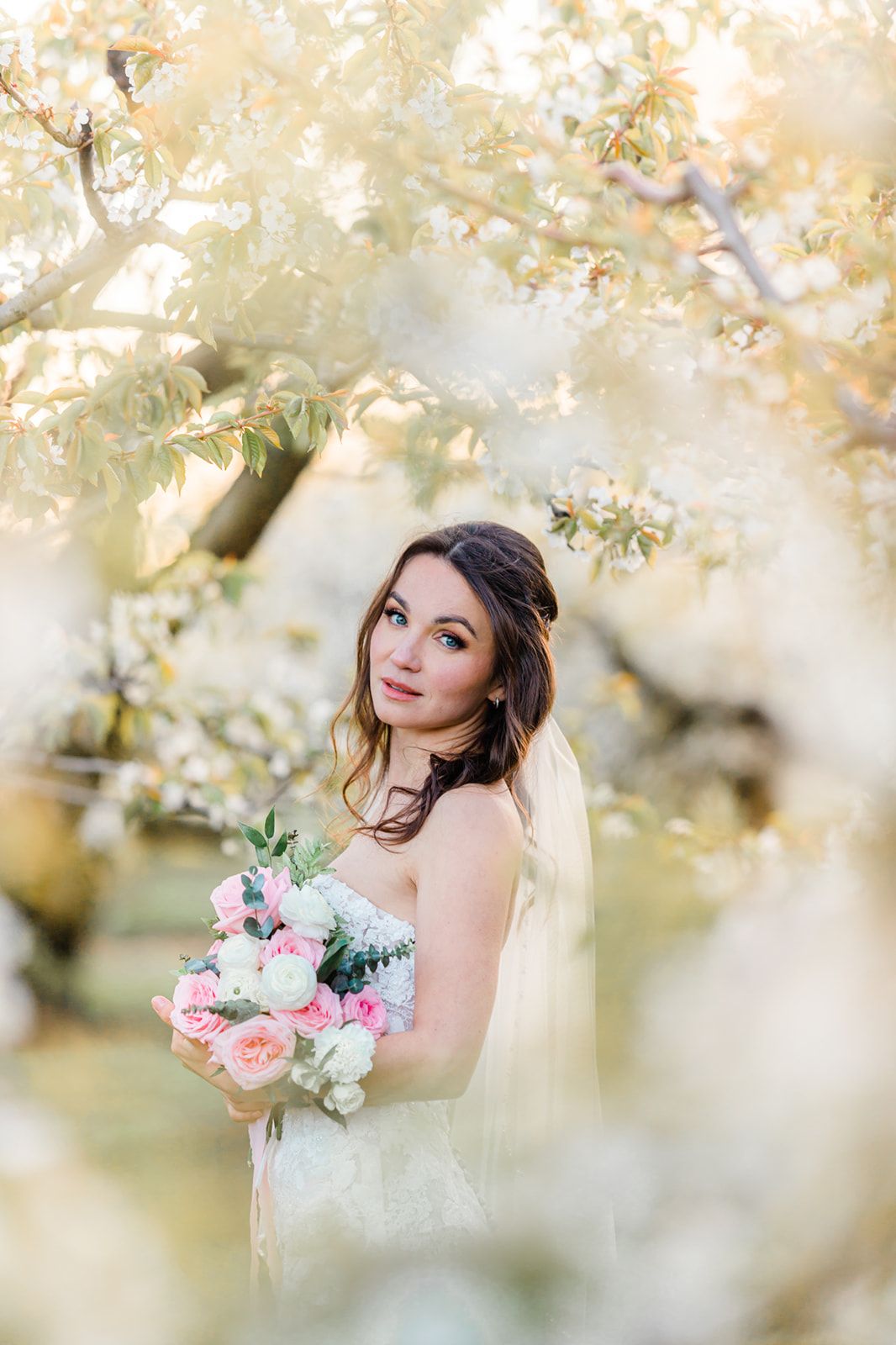 Elegant bridal portrait amidst blooming spring trees holding a pink and white bouquet.
