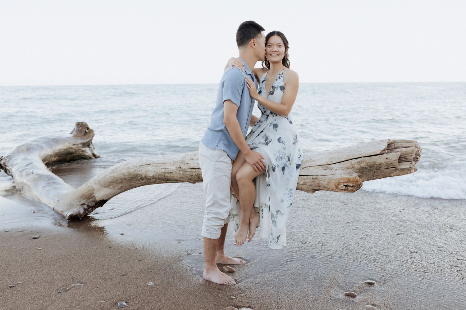 A couple poses romantically on driftwood at the beach with waves and horizon in the background.