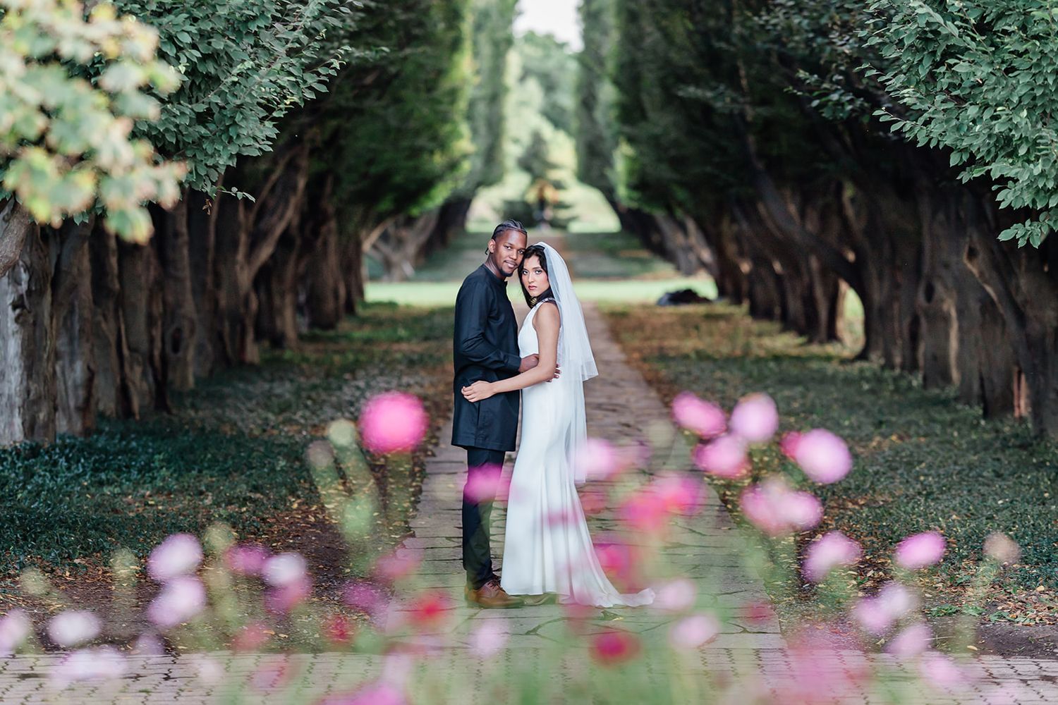 A romantic wedding photo of a couple embracing on a tree-lined path with pink flowers in the foreground.