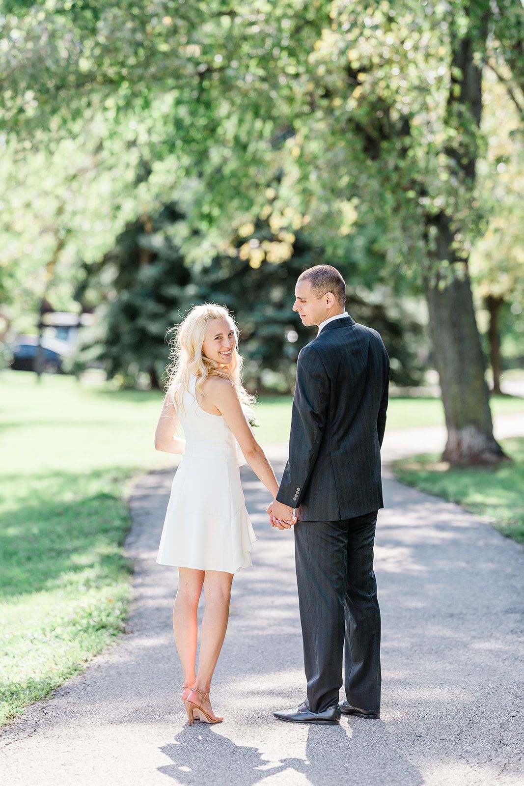 A couple in formal attire hold hands while standing on a sunlit path surrounded by green trees in a park.