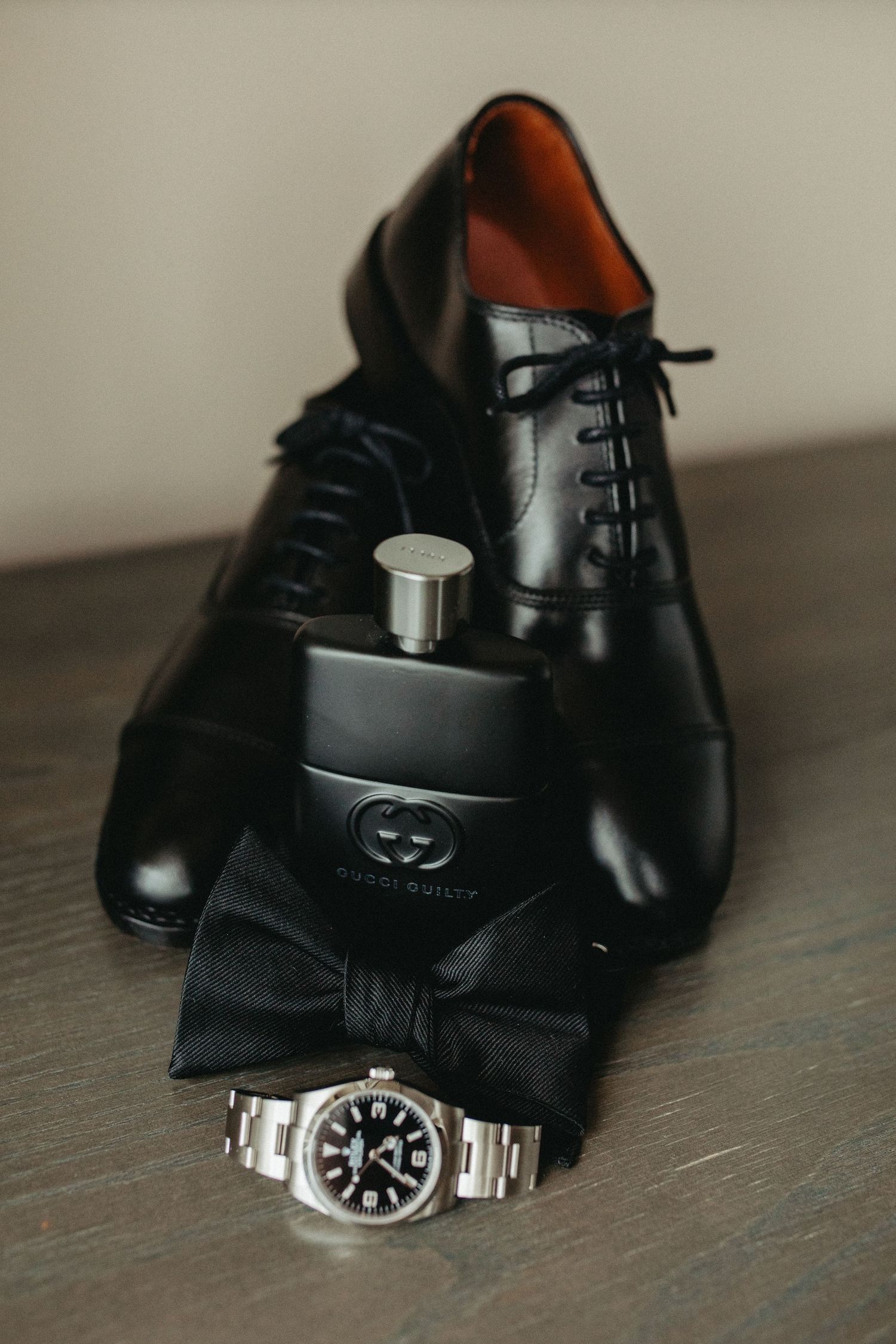 Black leather dress shoes with Gucci flask, bow tie and luxury watch arranged on wooden surface.