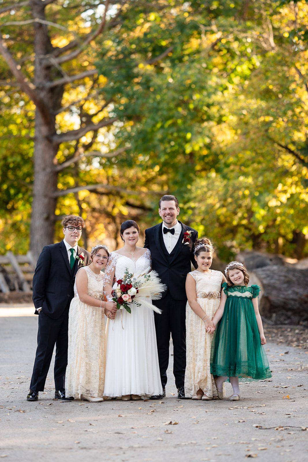 A wedding party stands outdoors on a dirt path surrounded by trees with golden autumn foliage in the background.