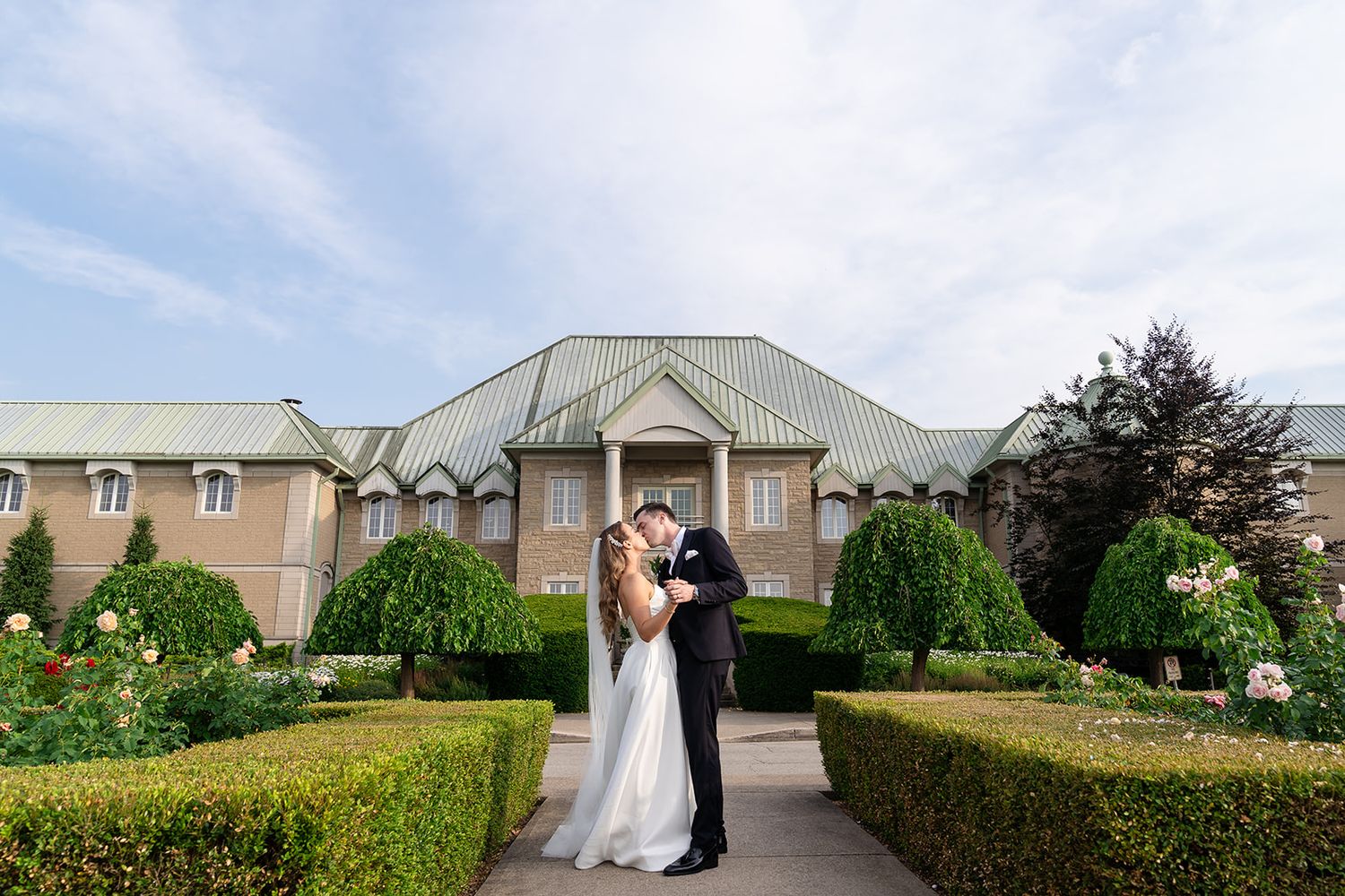 A romantic wedding couple stands in front of an elegant brick mansion with manicured gardens and hedges.