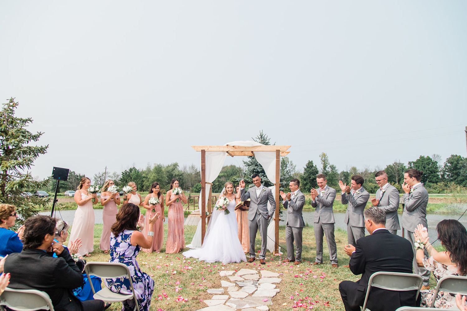 Outdoor wedding ceremony under wooden arch with bridal party in pink and grey standing on grassy field.