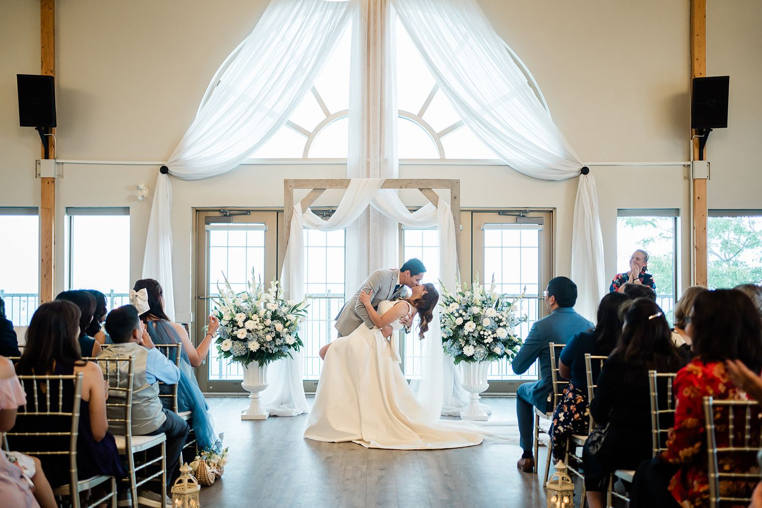An indoor wedding ceremony in a bright venue with white drapes and floral arrangements.