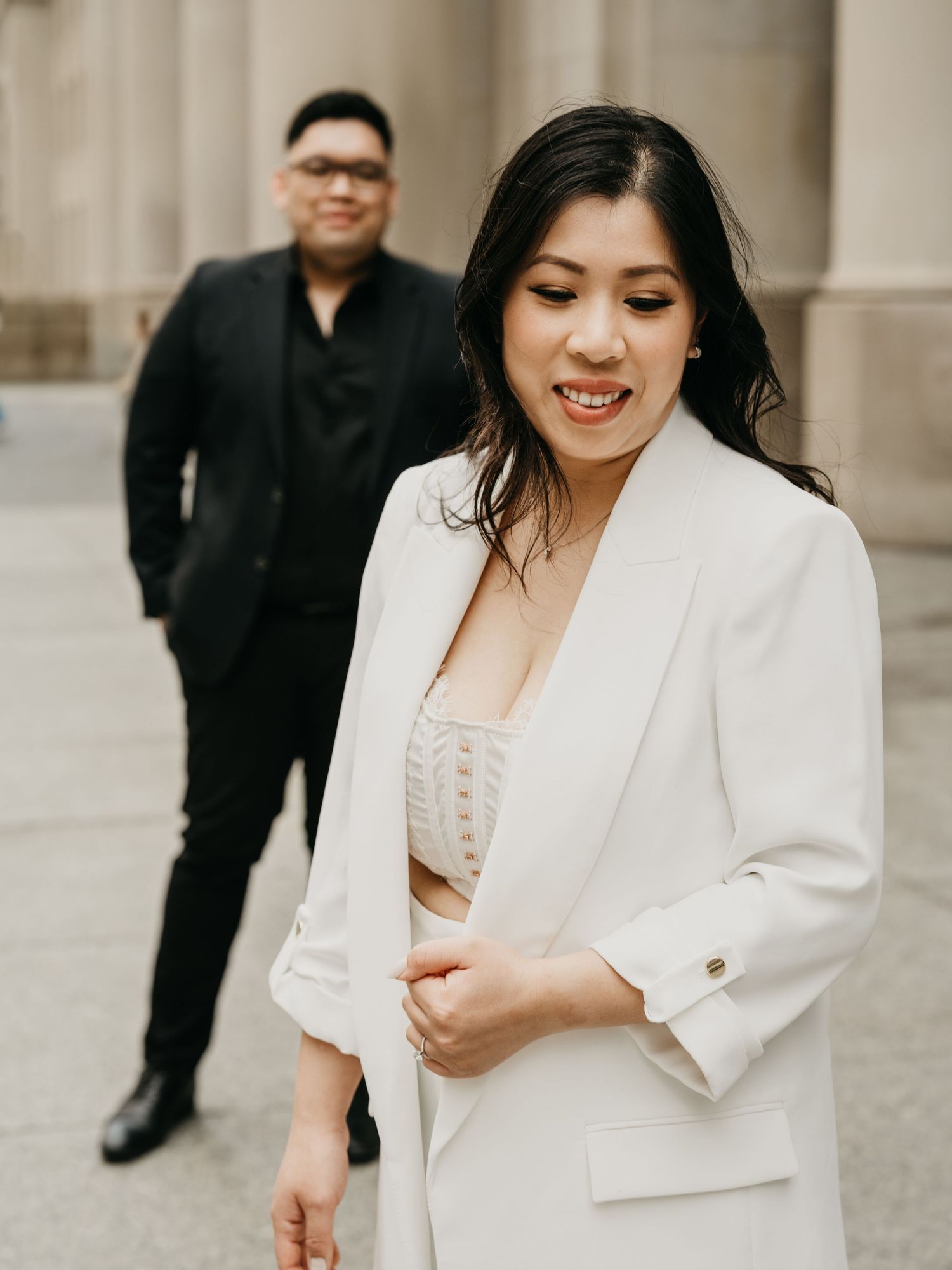 A couple in formal attire poses outside a building with classical columns, with someone in white in front.