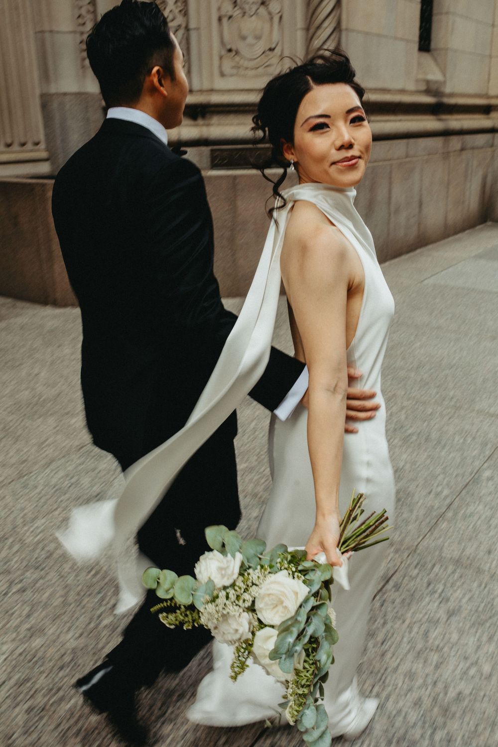 Elegant wedding couple walks together as bride holds a delicate white and green bouquet against classical architecture.