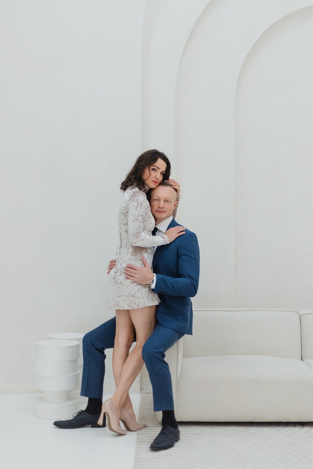 A couple in formal attire embraces on a white couch against a minimalist white backdrop.
