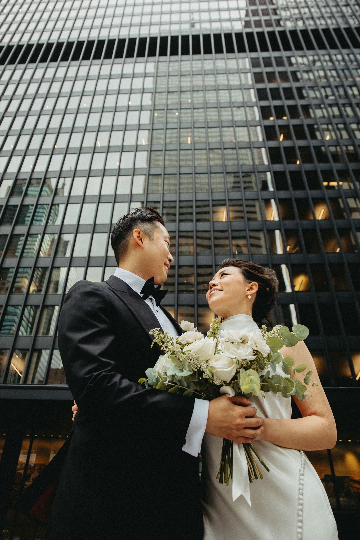 A couple in wedding attire embrace while holding a white floral bouquet in front of a modern glass skyscraper.
