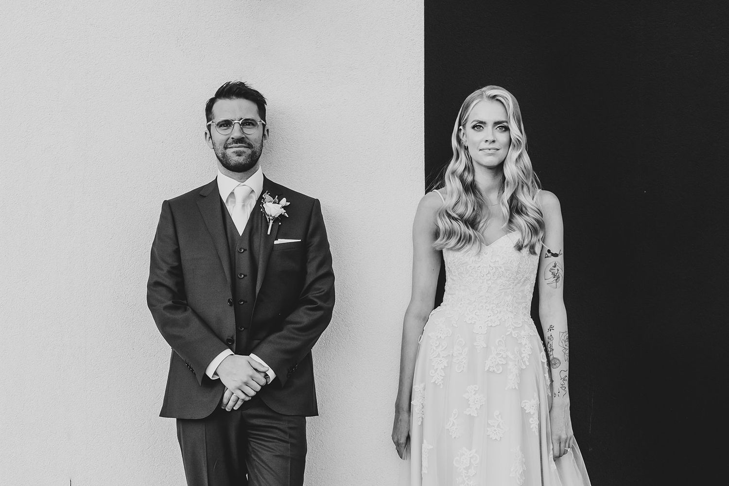 A black and white wedding portrait of a bride in a white gown and groom in dark suit standing against a wall.