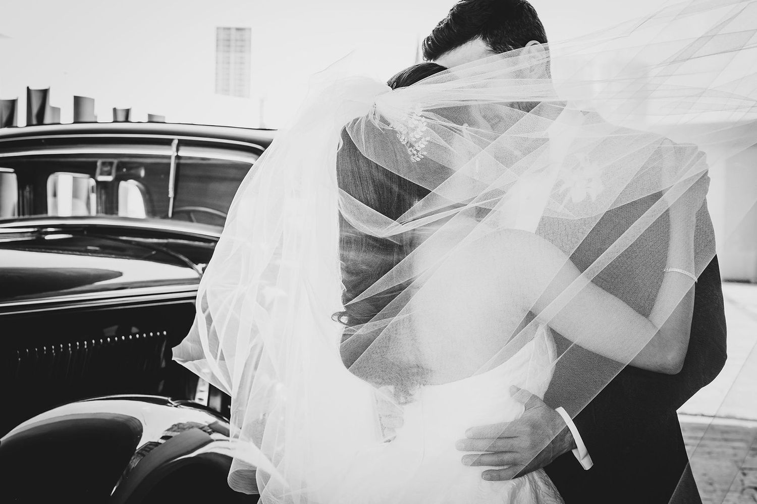 A classic black and white wedding photo with flowing veil and vintage car in the background.