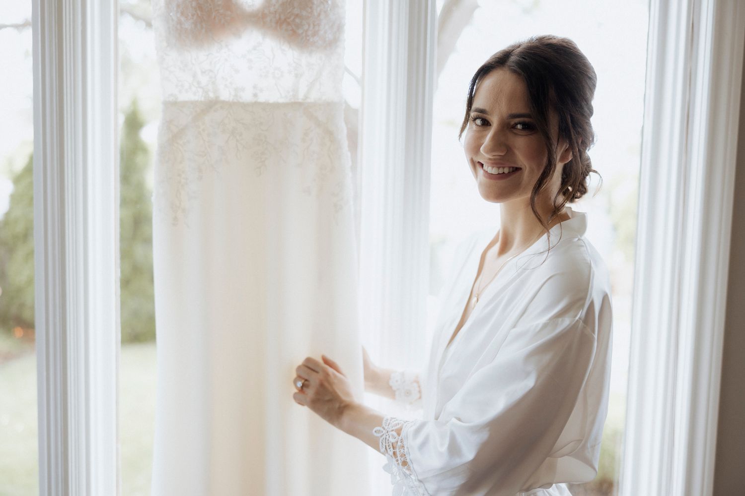 A person in a white robe stands by a bright window next to a hanging wedding dress.