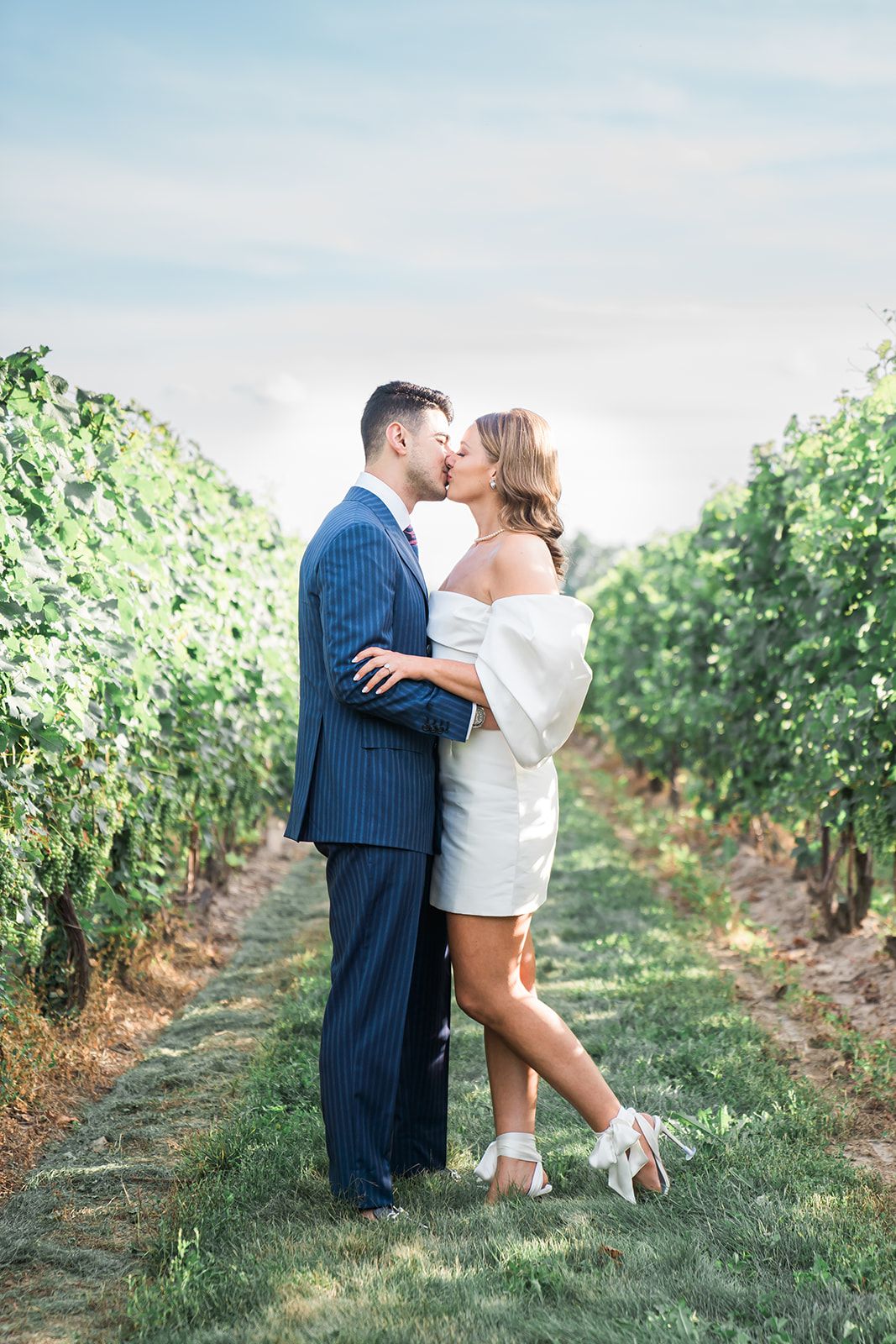 Romantic couple embraces in vineyard setting wearing formal attire on sunny afternoon.