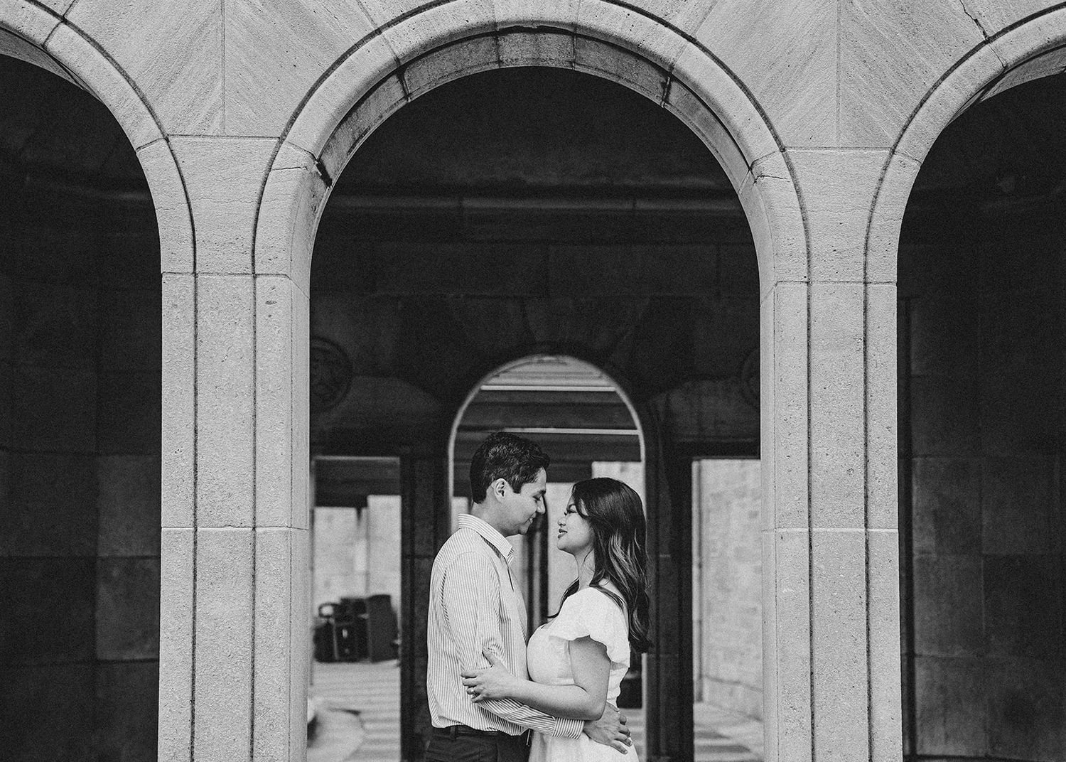 Romantic couple embracing under a classic stone archway in black and white.