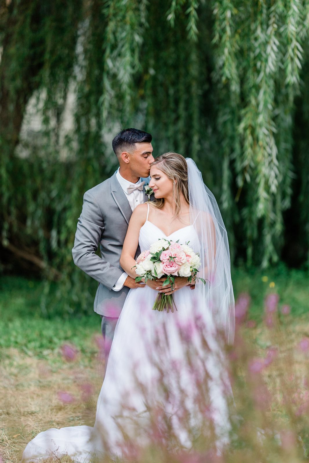Romantic wedding couple embracing under cascading willow trees with pink florals.