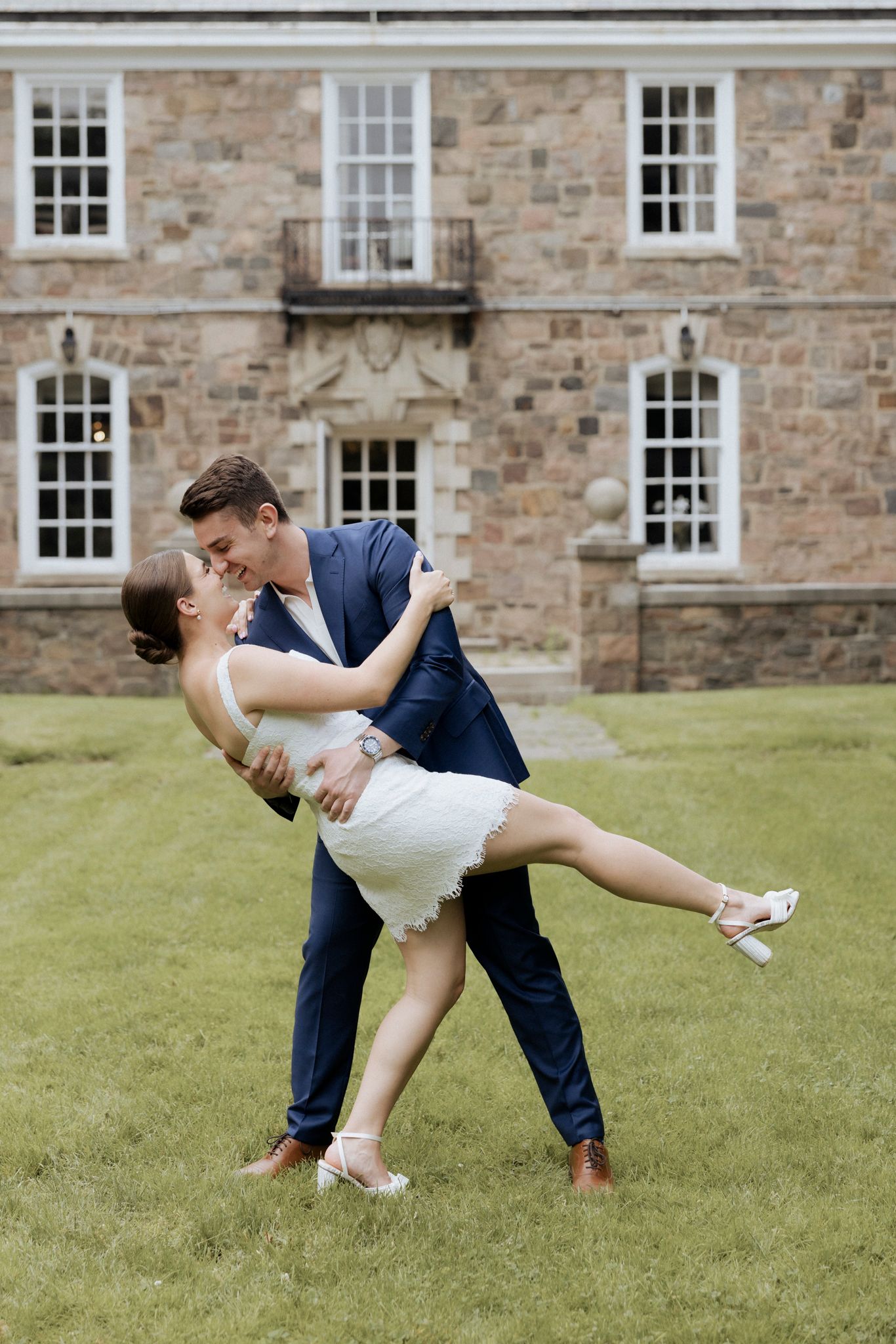 A romantic couple dances and embraces on a lawn in front of a stone building with white windows.