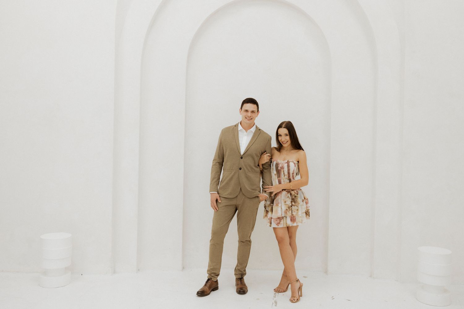 A couple in formal attire poses together against a white arched wall background.